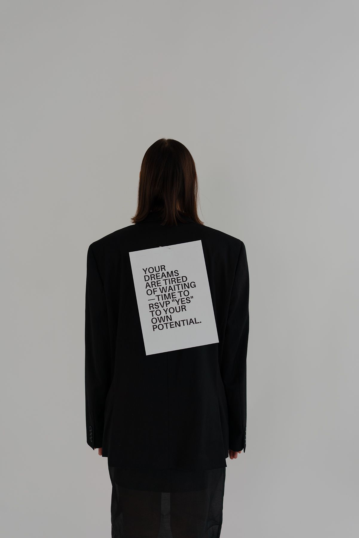 A person wearing a black blazer with white text on the back stands against a light gray background.