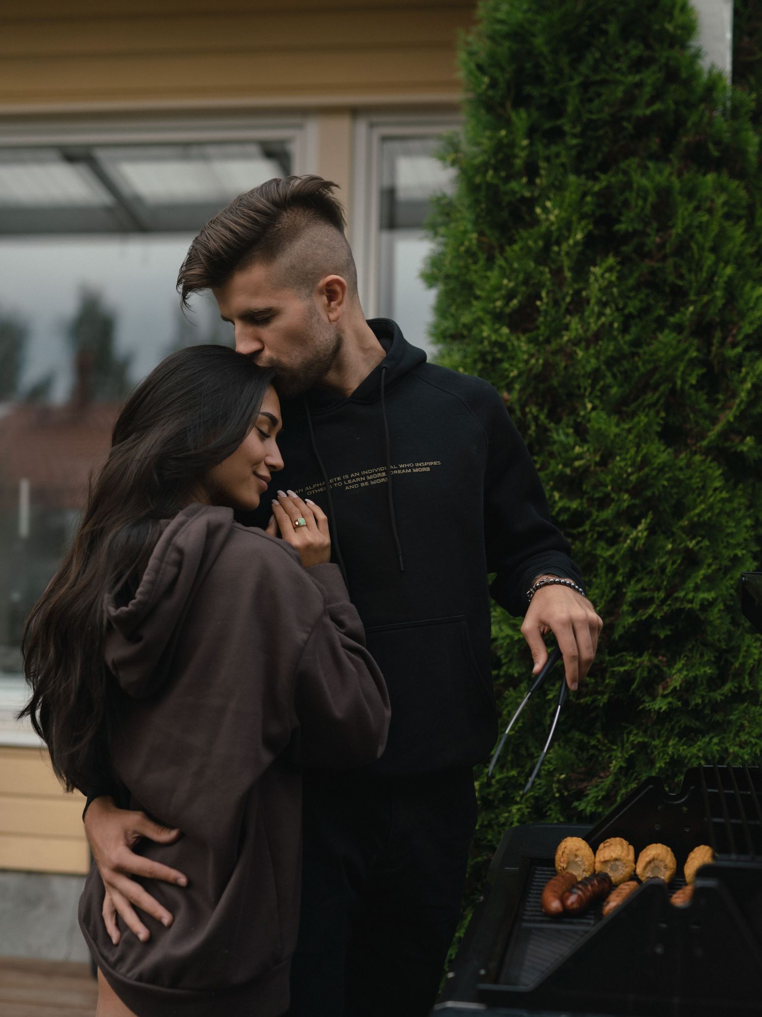 A couple shares an intimate moment outdoors by evergreen trees near a barbecue grill.