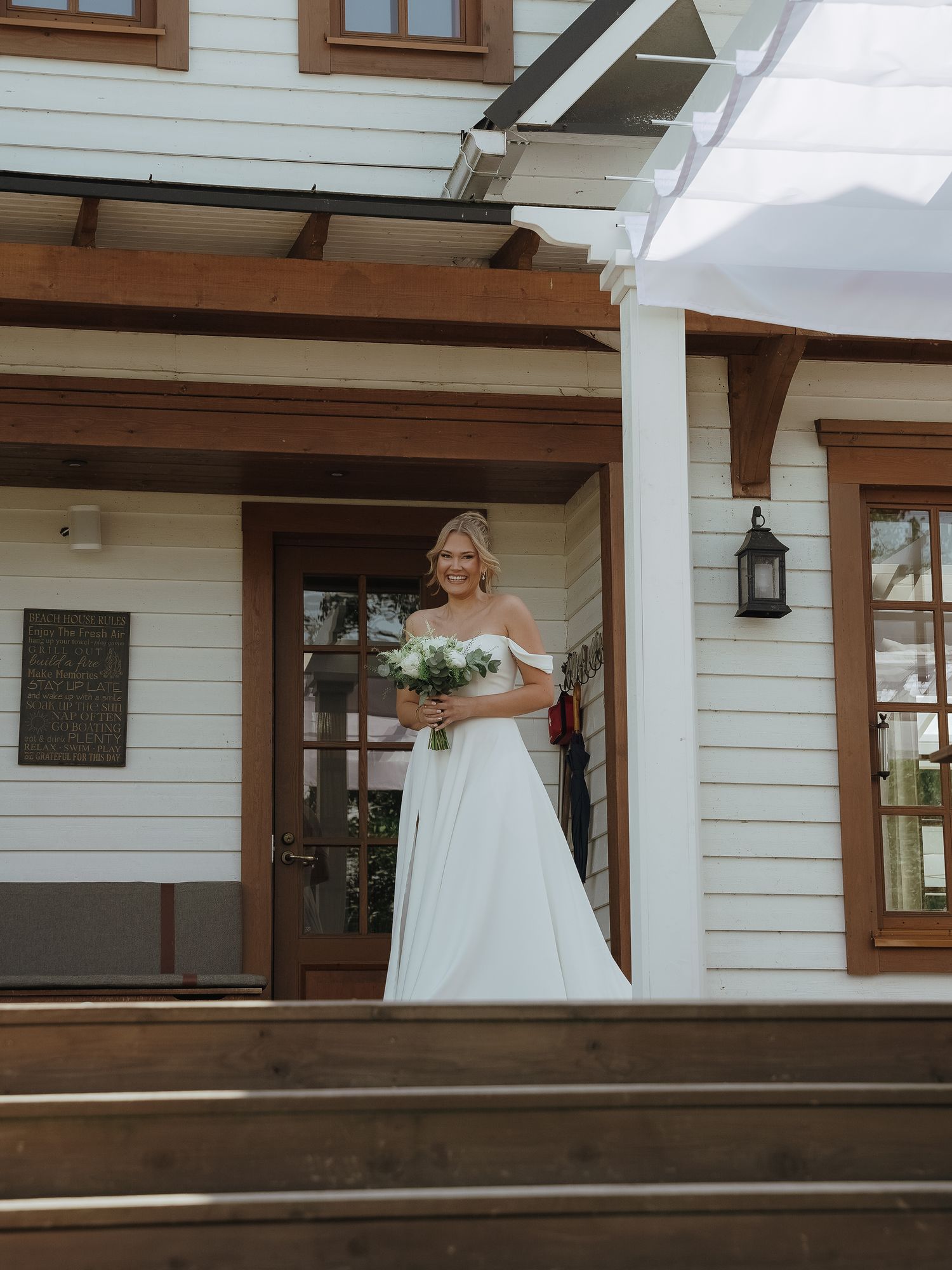 A bride in a white off-shoulder wedding dress stands on the front porch steps of a white farmhouse-style home.