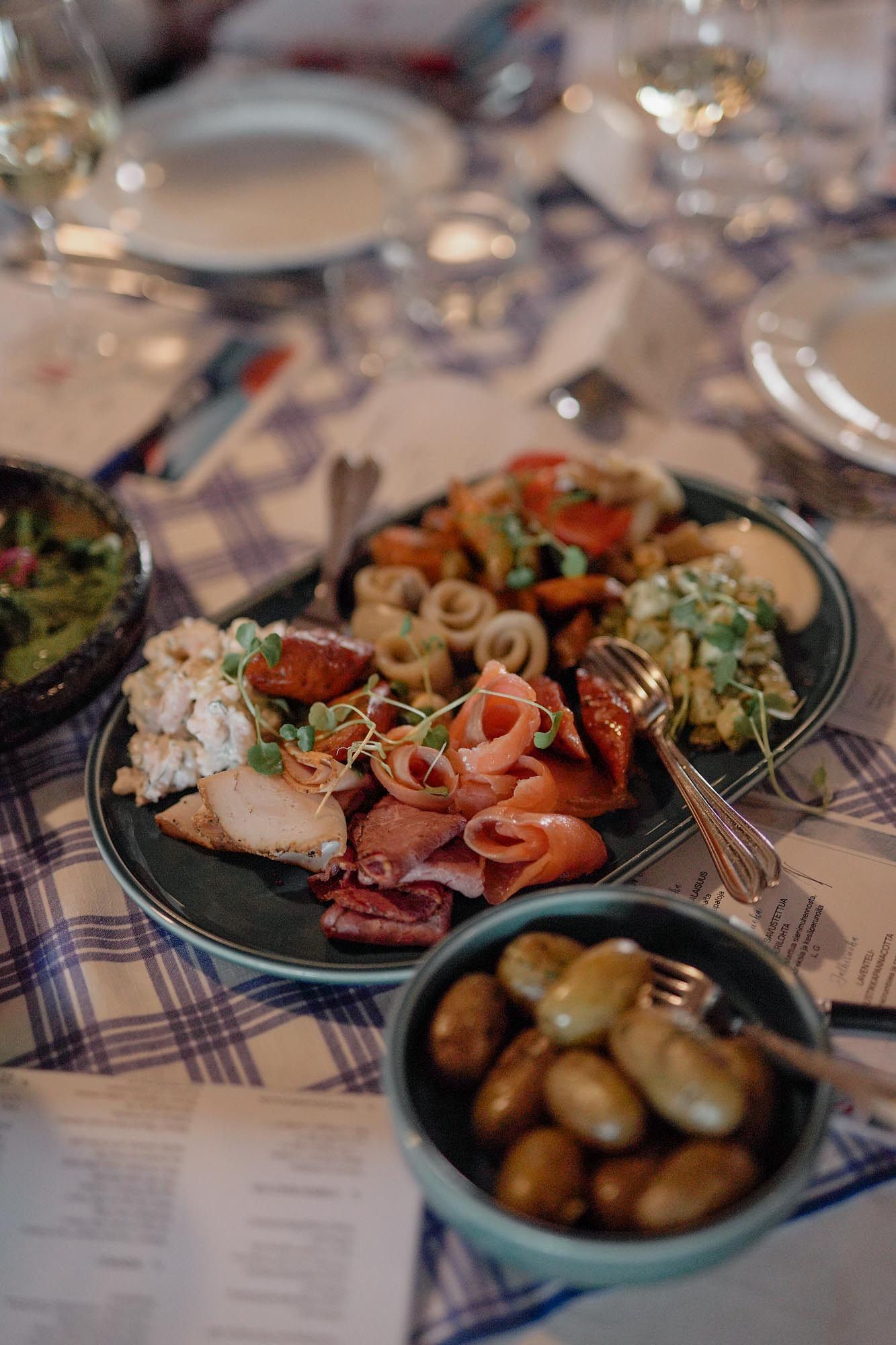 Elegant charcuterie platter with salmon, meats, calamari, and microgreens beside a bowl of marinated olives on a plaid tablecloth.