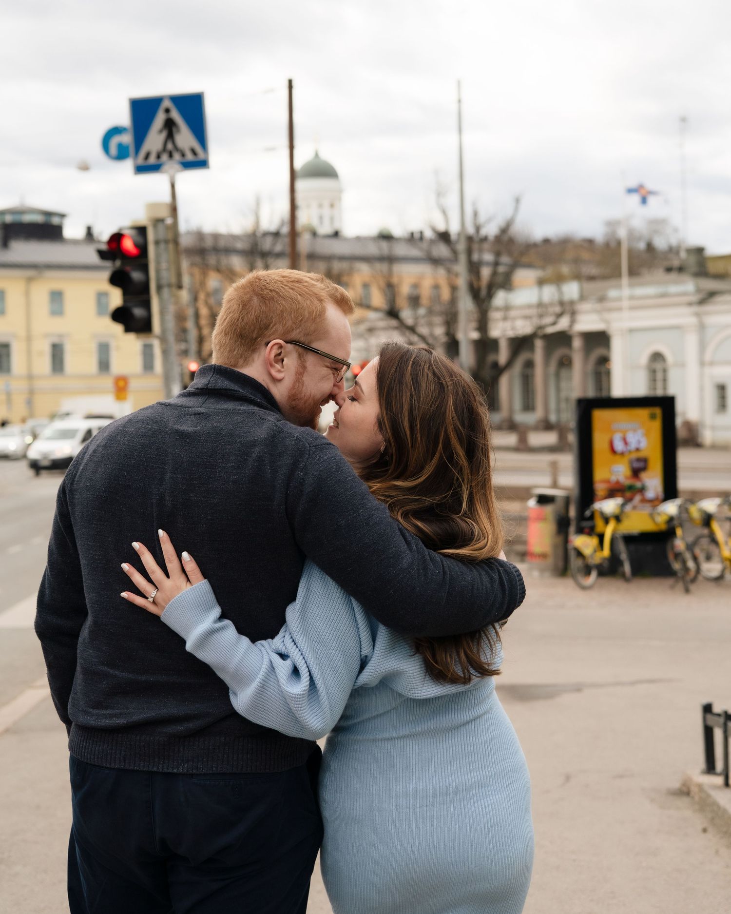Couple sharing a romantic kiss on a city street corner with traffic lights and historic building in background.
