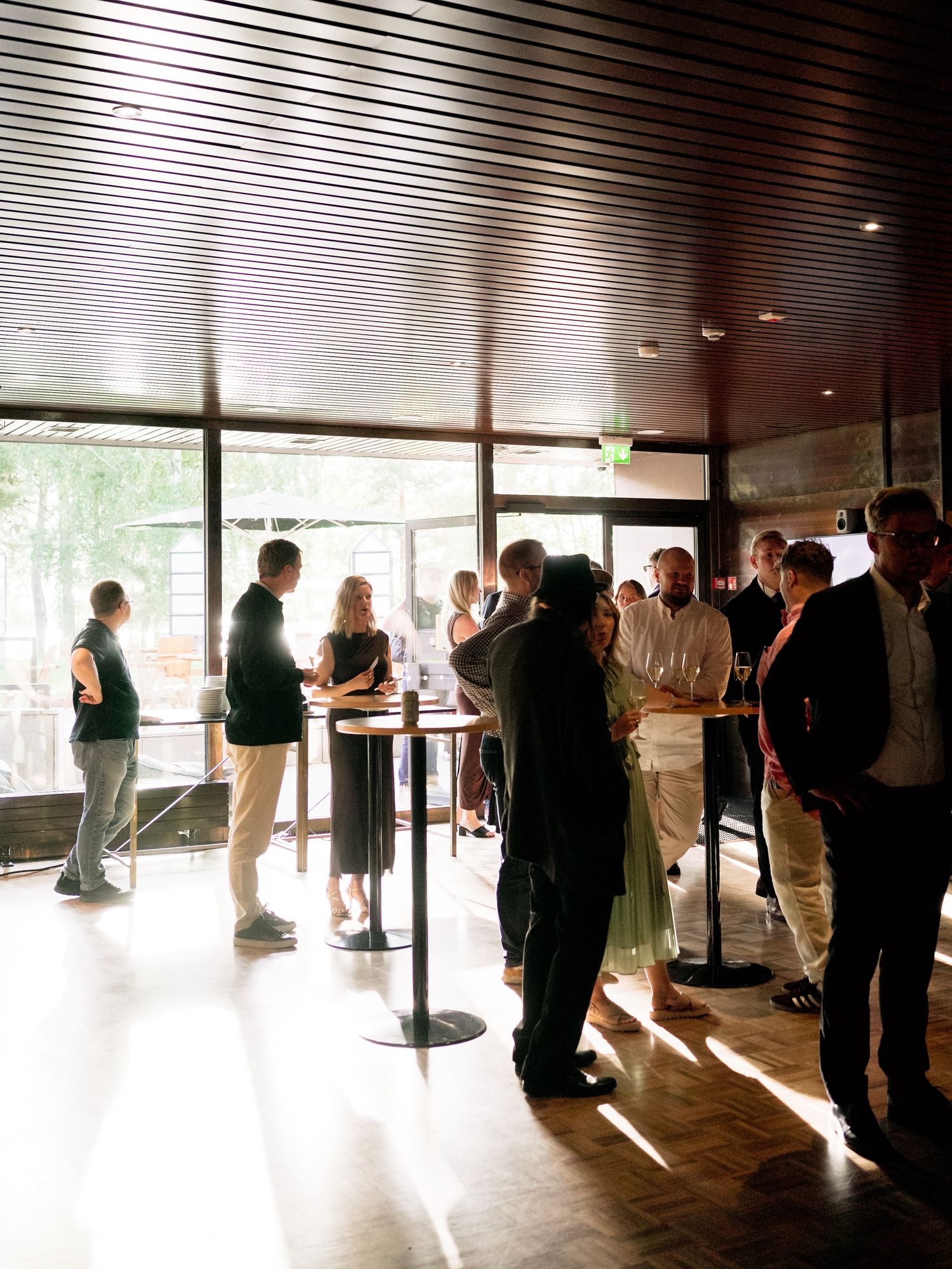 Guests mingle at an elegant indoor cocktail reception with large windows and warm sunlight flooding the venue.