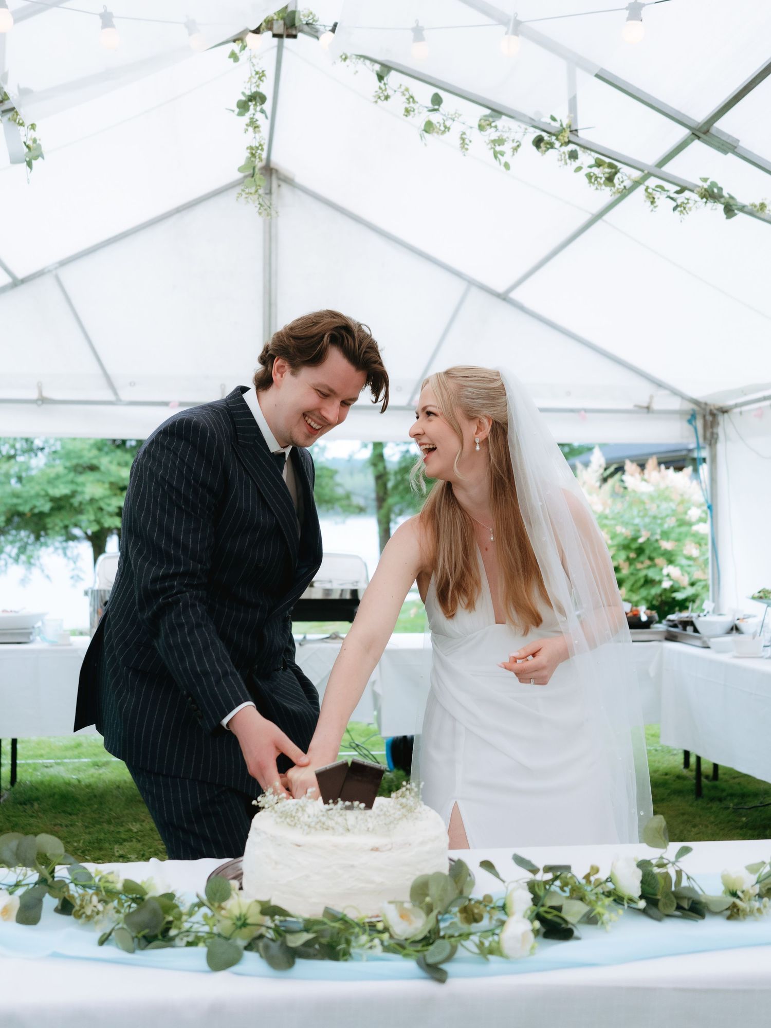 Bride and groom smiling while cutting white wedding cake under elegant tent with floral greenery decor.
