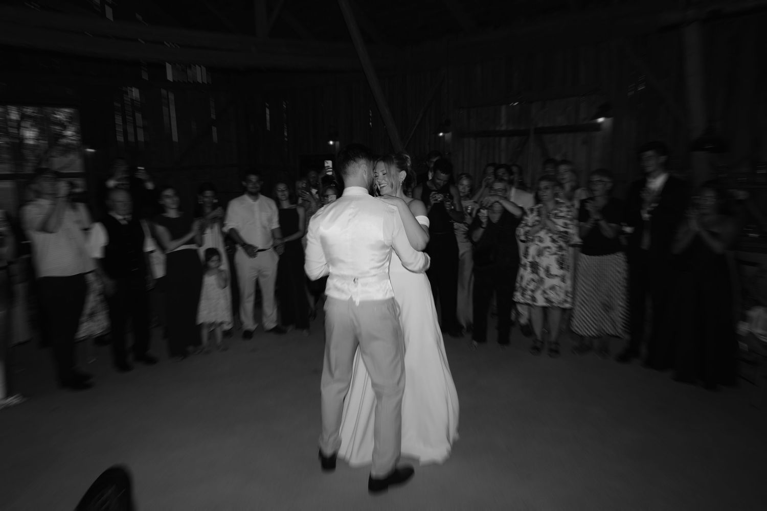 A black and white photo of a wedding couple sharing their first dance surrounded by guests on the dance floor.