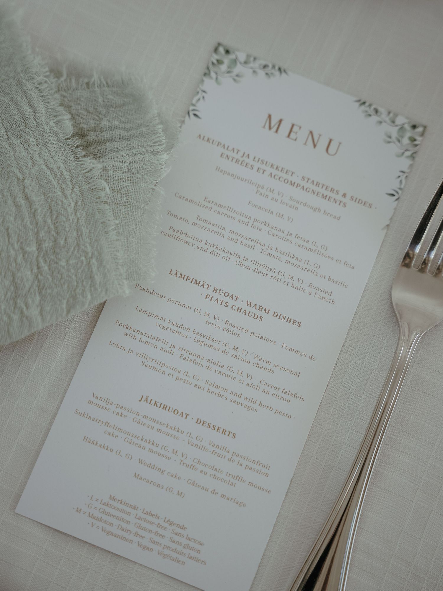 Elegant white wedding menu card with decorative borders displayed next to silverware on a light colored surface.