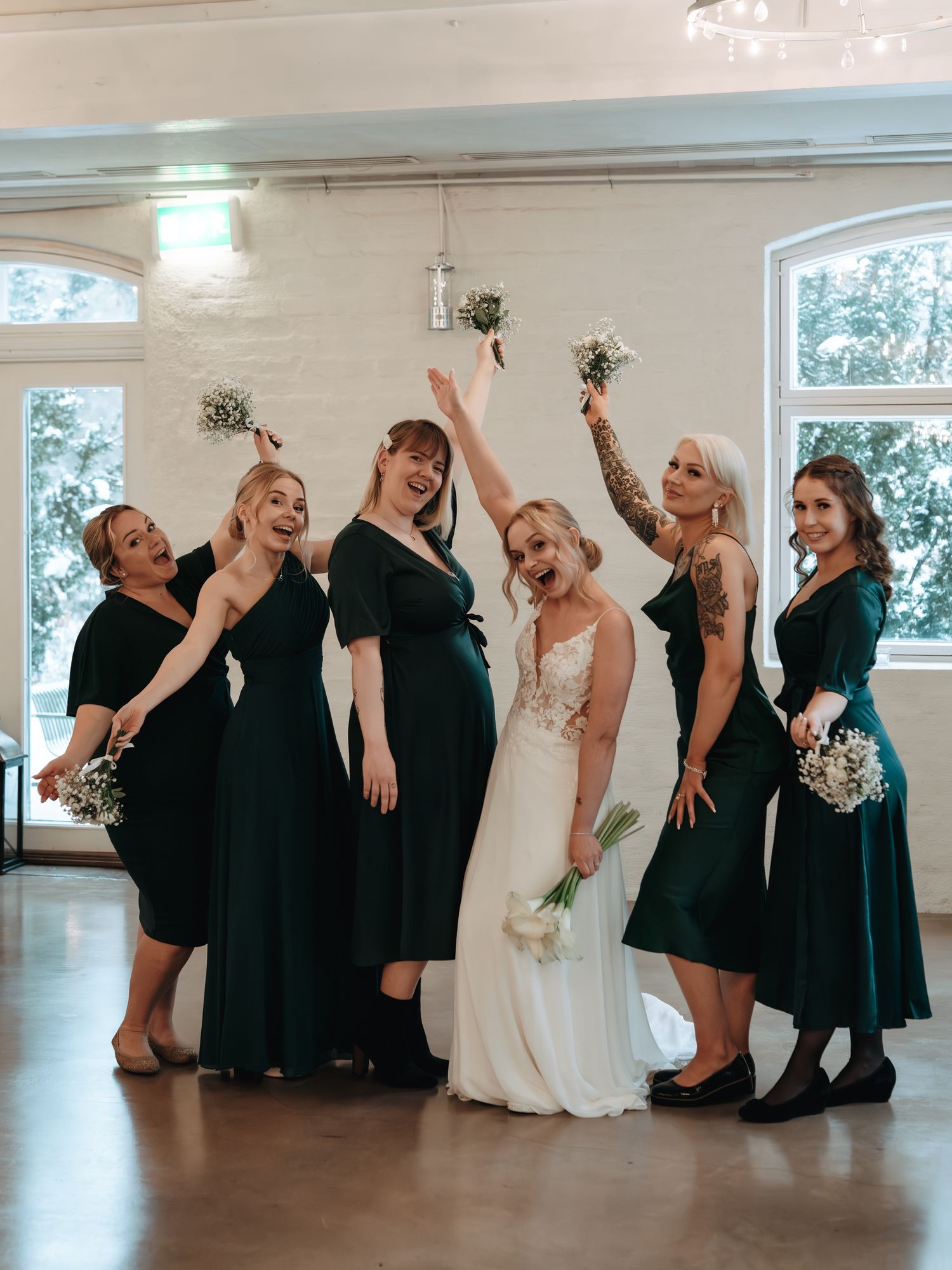 Wedding party in dark green bridesmaid dresses and white bridal gown pose playfully together in a bright indoor venue.