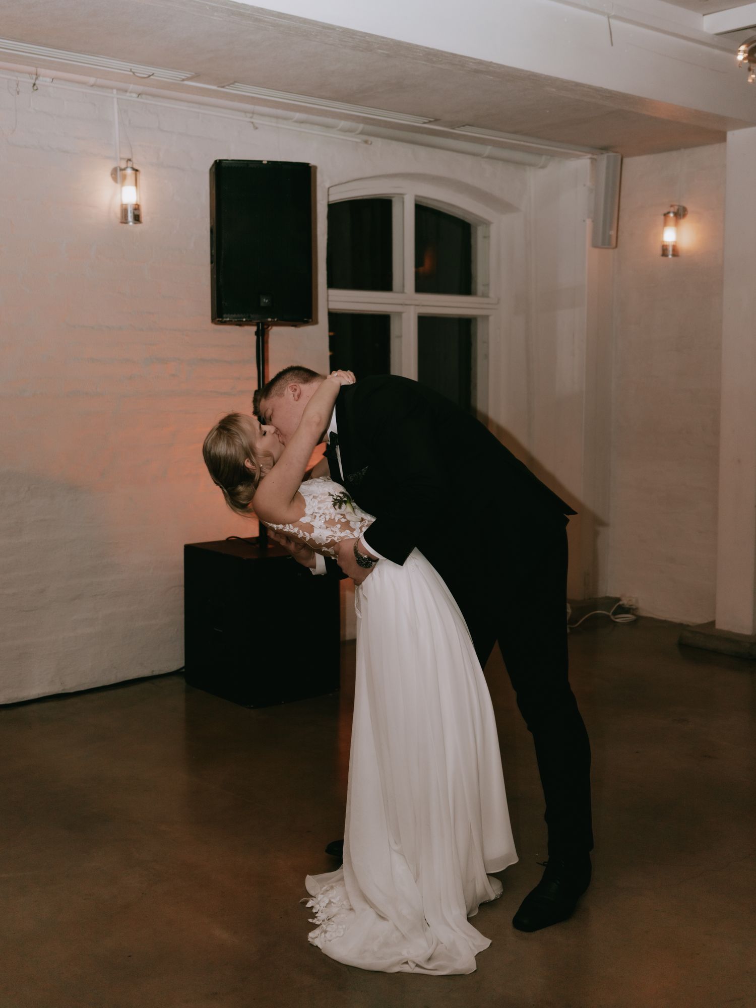 A couple shares a romantic dance movement in a white-walled room with sconce lighting and a black upright piano.