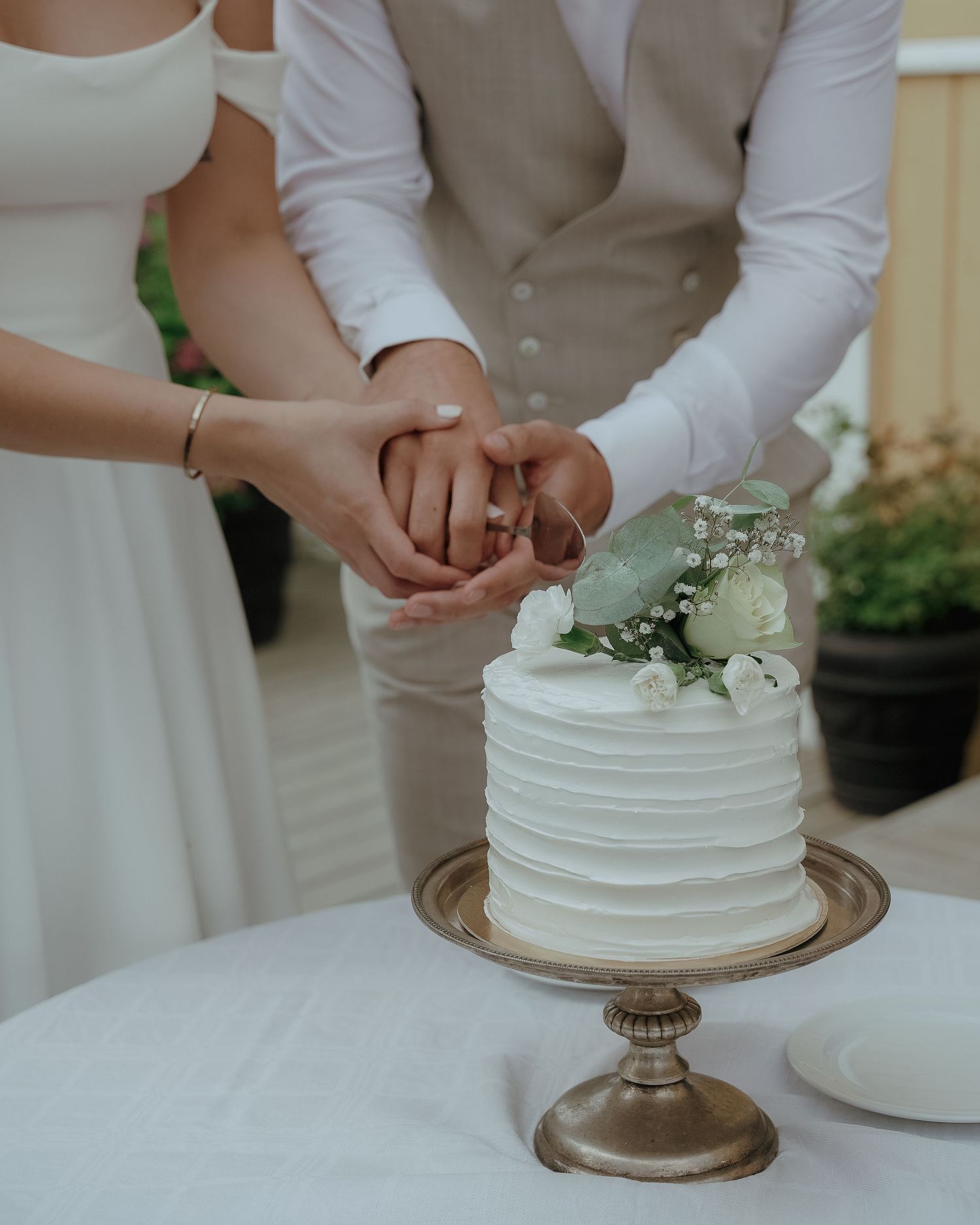 Couple's hands cutting into an elegant white wedding cake with floral decoration atop a gold pedestal stand.
