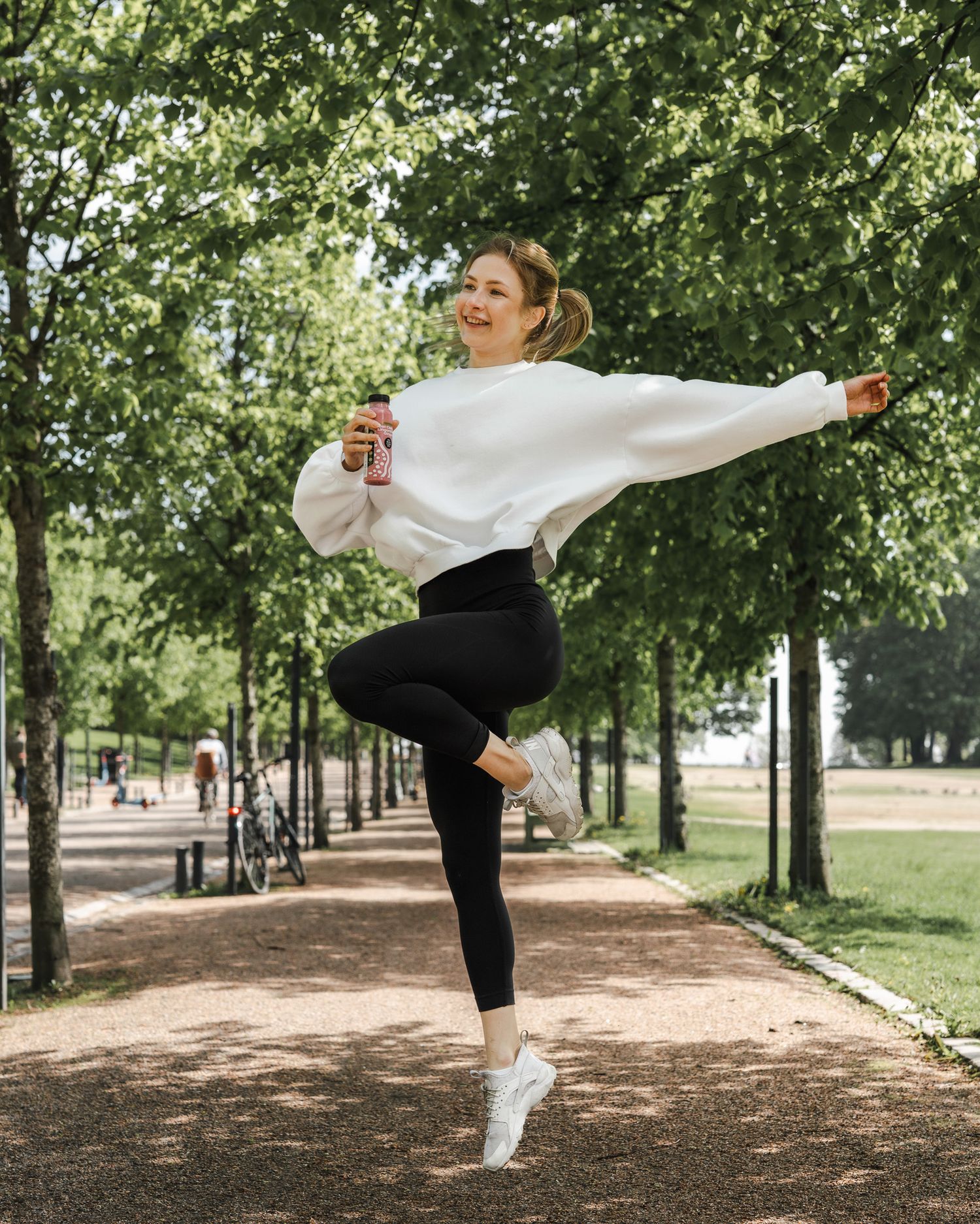 Someone in a white sweater and black leggings joyfully dances on a tree-lined path while holding a beverage.