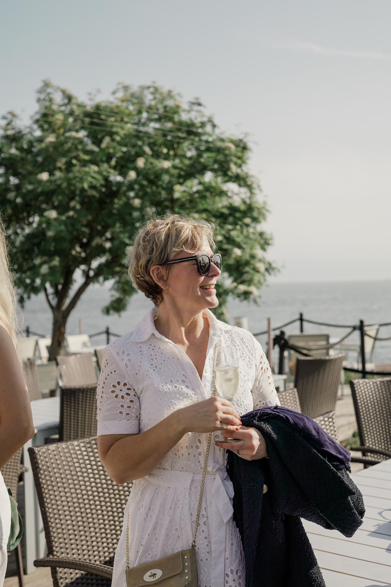 Smiling woman in white dress and sunglasses holding champagne glass on sunny outdoor terrace by the sea.