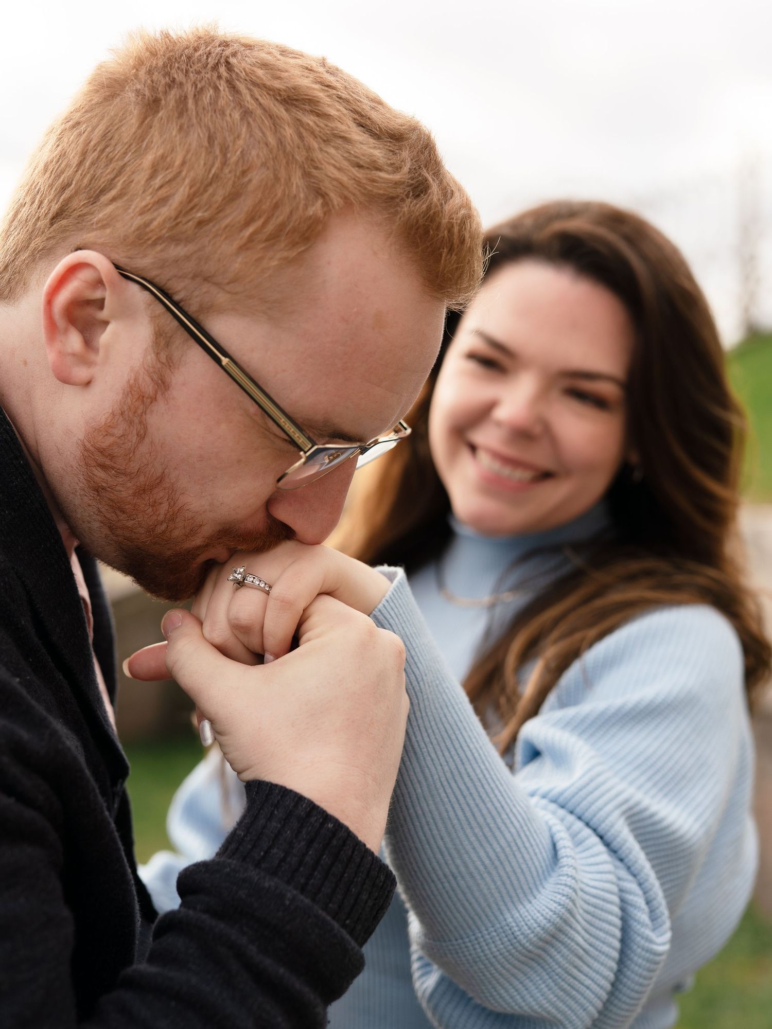 A red-haired man with glasses kisses a woman's hand while she smiles warmly during an outdoor engagement photo.