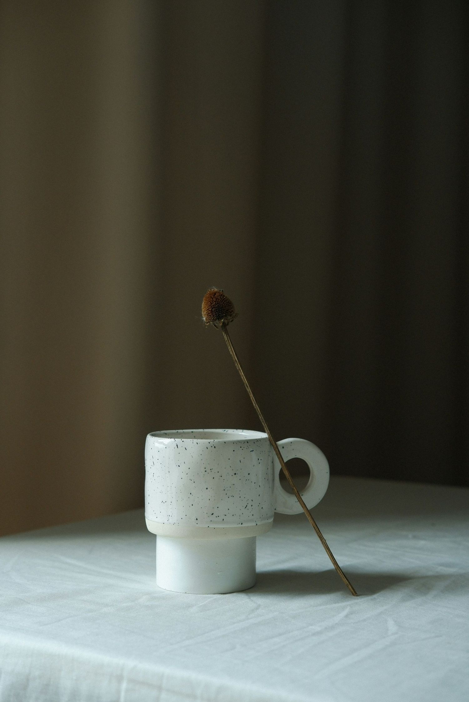 A minimalist white ceramic coffee mug sits on a light tablecloth with a single dried flower stem leaning against it.