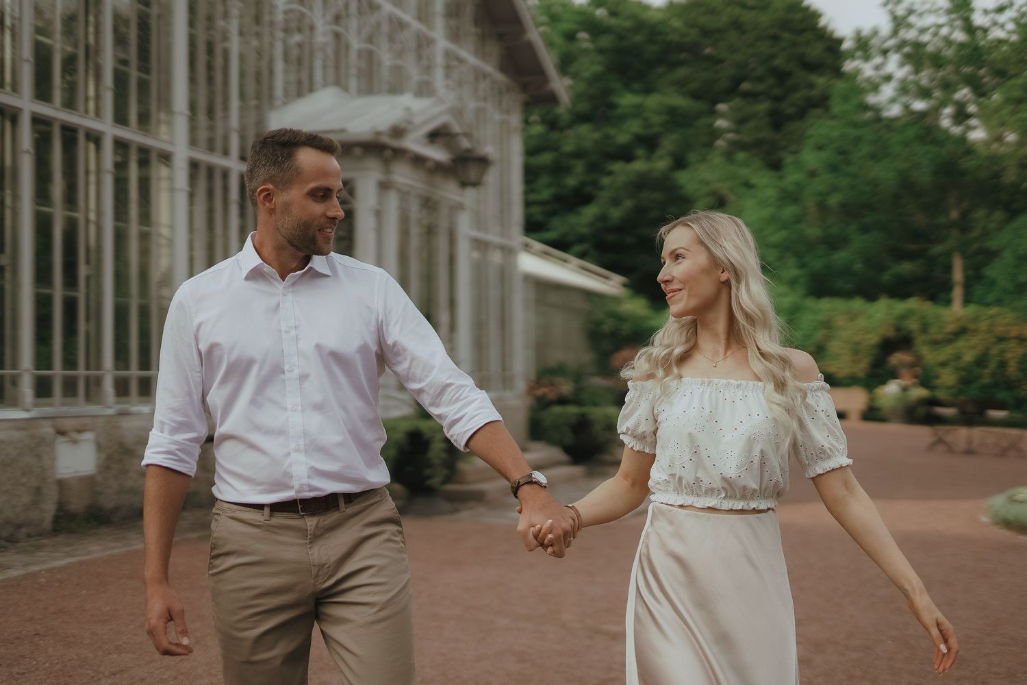 A romantic couple holding hands and walking together near a glass conservatory in casual summer attire.