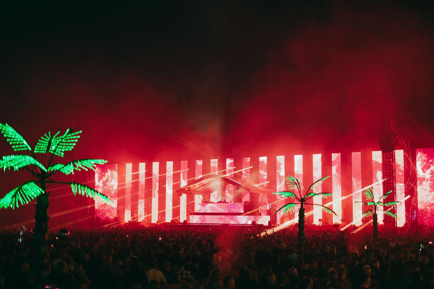 Large crowd at outdoor music festival with neon palm trees and red lighting effects illuminating a stage display.