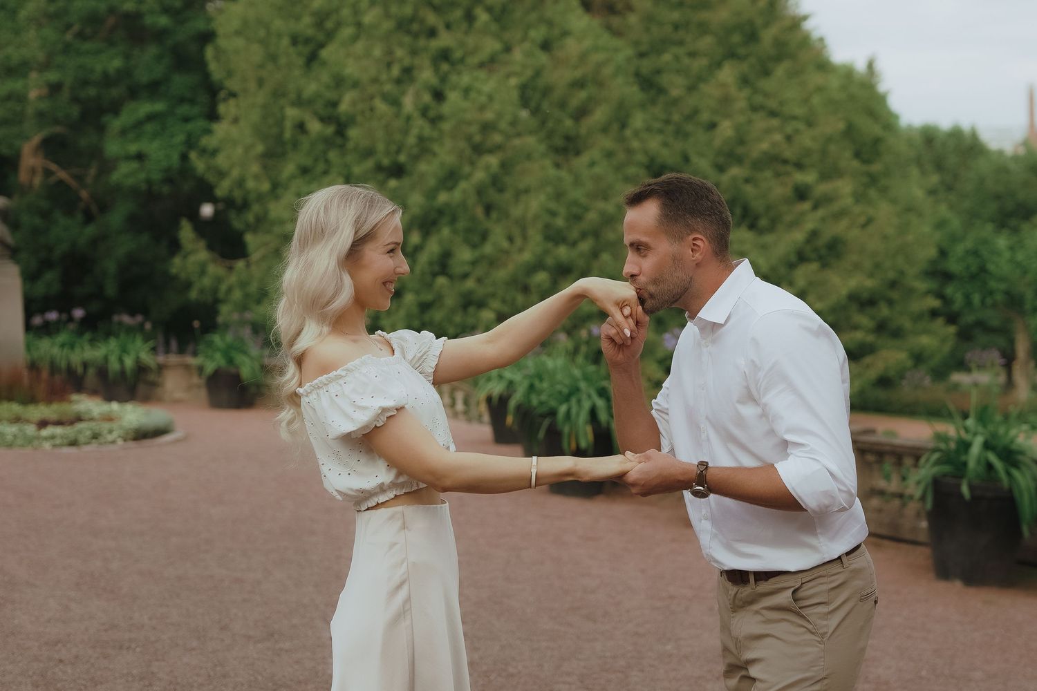 A couple in a garden path celebrates joyfully as someone kisses another's hand in a romantic moment.