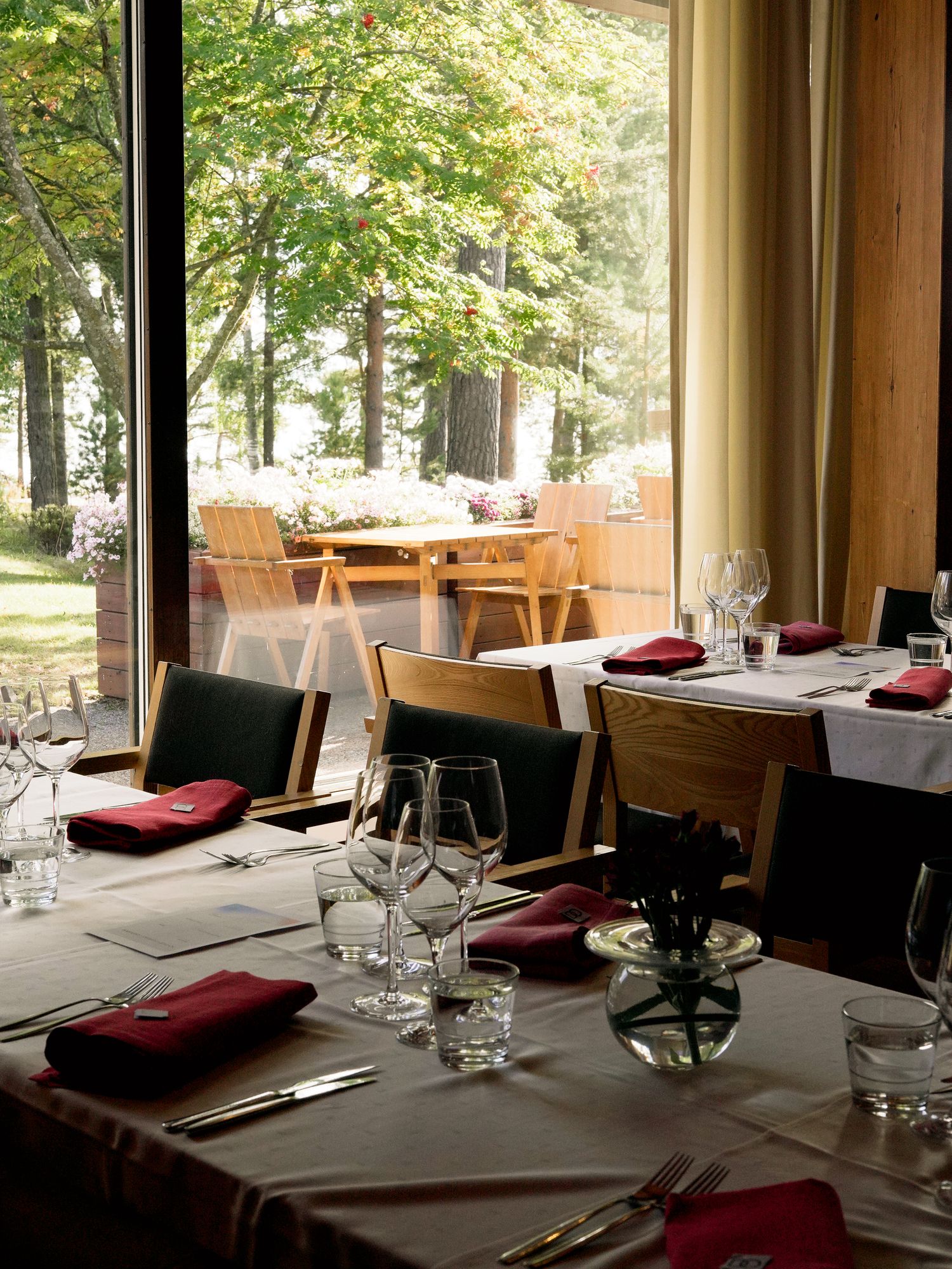 Elegant restaurant dining room with red napkins, wine glasses, and large windows overlooking a lush green tree-lined patio.