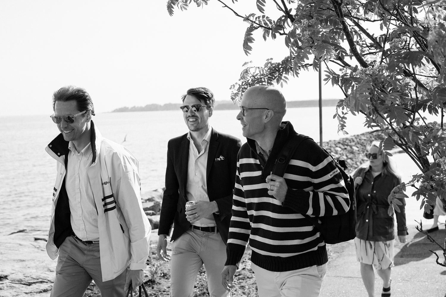 Three men walking along a waterfront path in black and white photo, dressed in smart casual attire.
