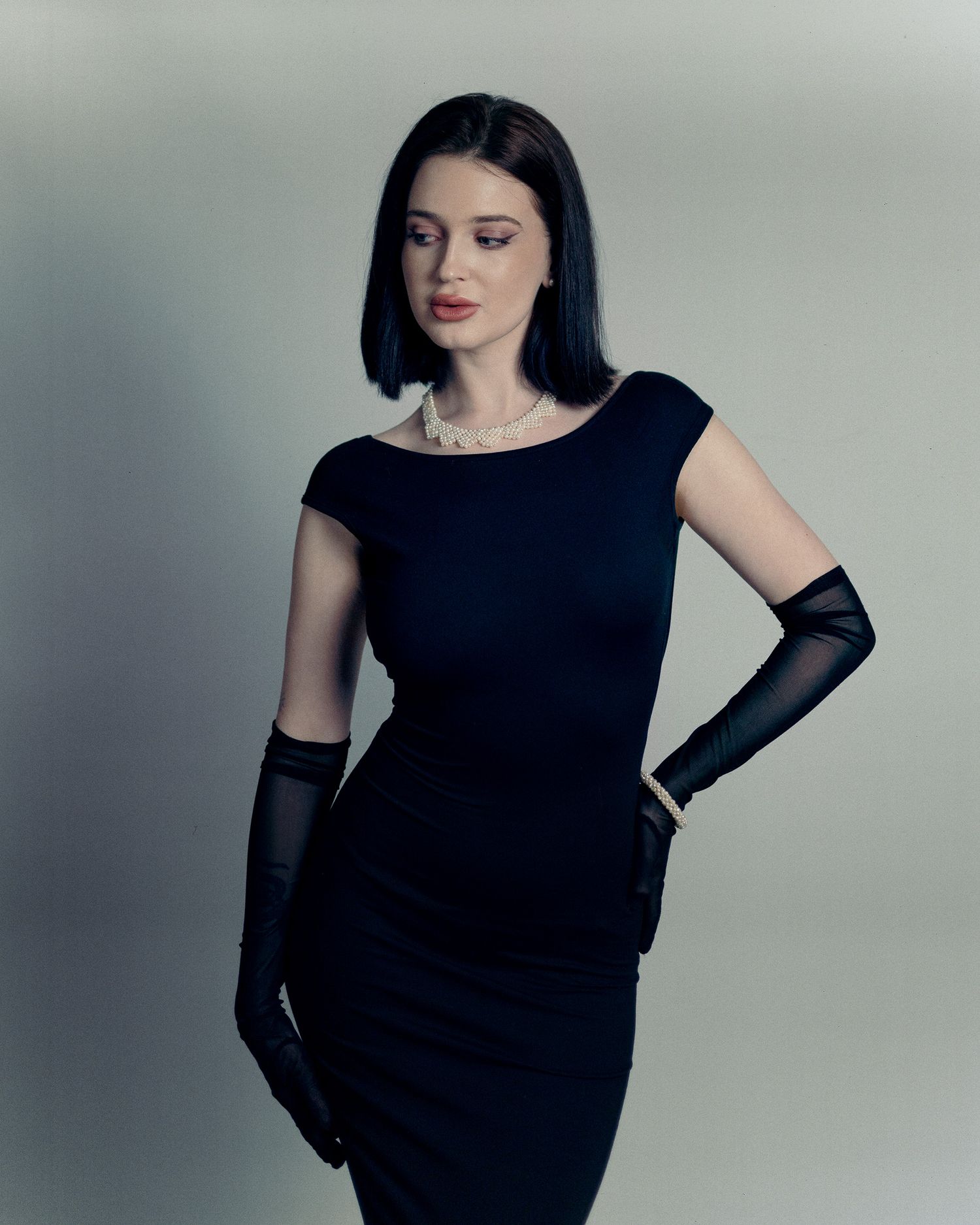 Classic fashion pose showing black evening dress with elbow-length gloves against light gray background.