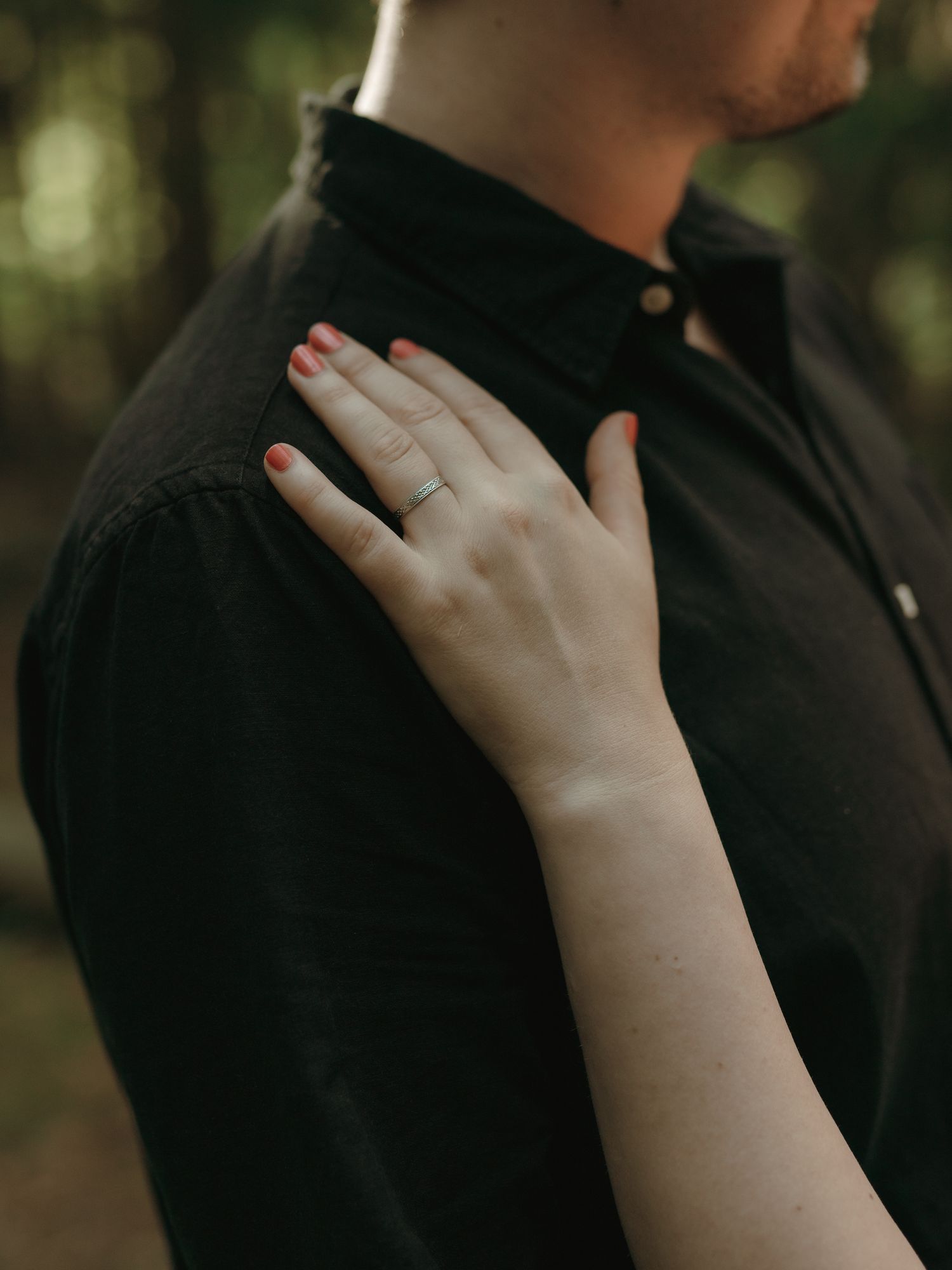 A delicate hand with red painted nails rests tenderly on a shoulder wearing a black button-up shirt in a romantic outdoor setting.