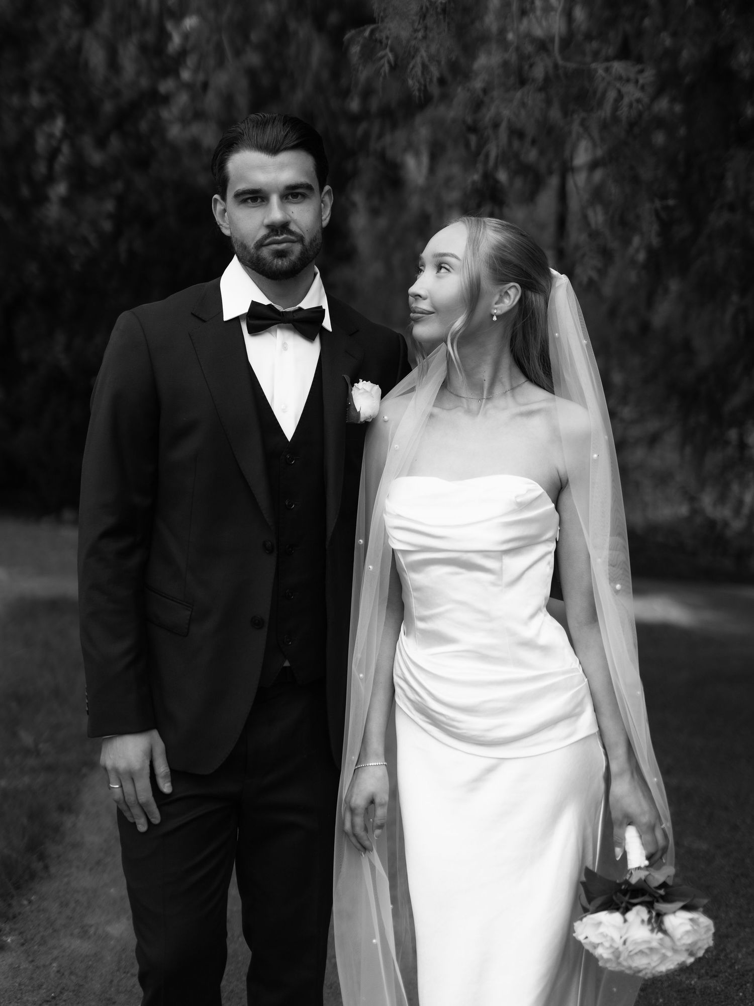 Black and white wedding photo of bride in strapless gown with veil and groom in tuxedo walking outdoors.