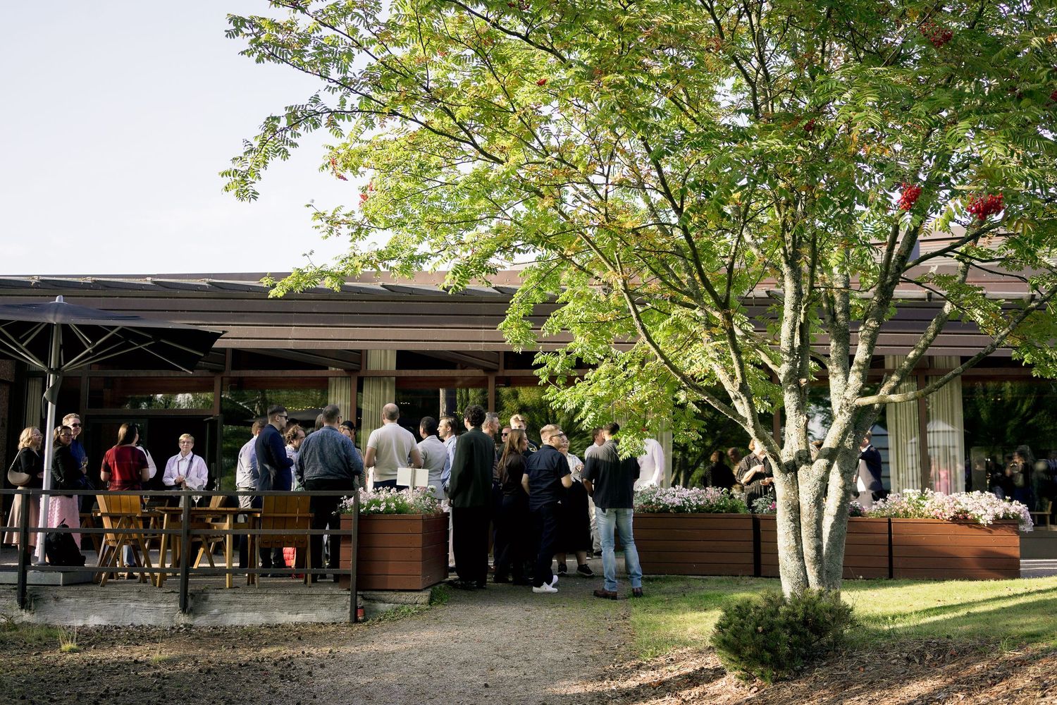 Guests mingle outdoors at a restaurant patio event surrounded by lush trees and wooden planters.
