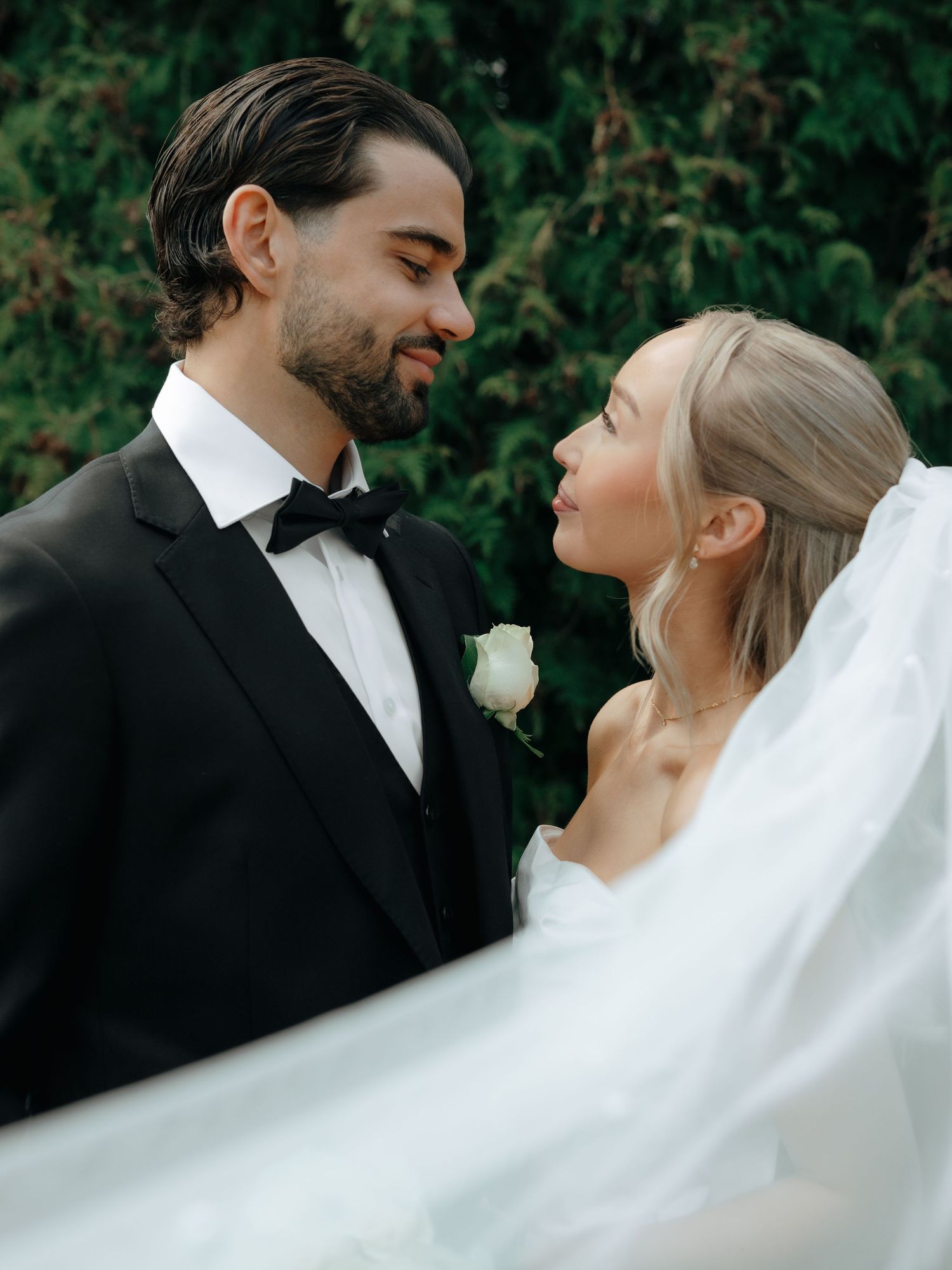 A bride and groom in formal wedding attire share a romantic moment outdoors, with a flowing veil and lush greenery behind them.