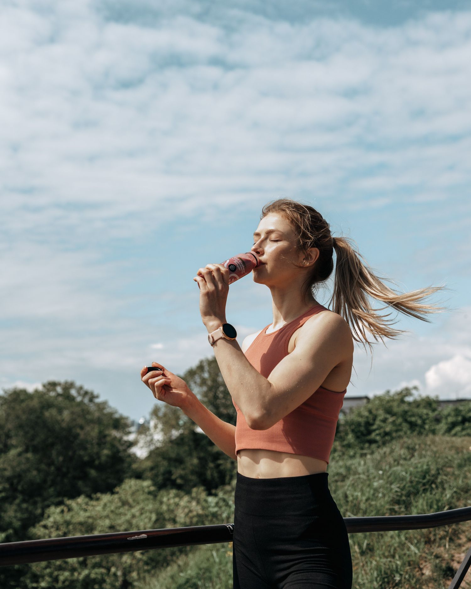 A fit person in athletic wear drinks from a water bottle outdoors on a sunny day with trees in the background.