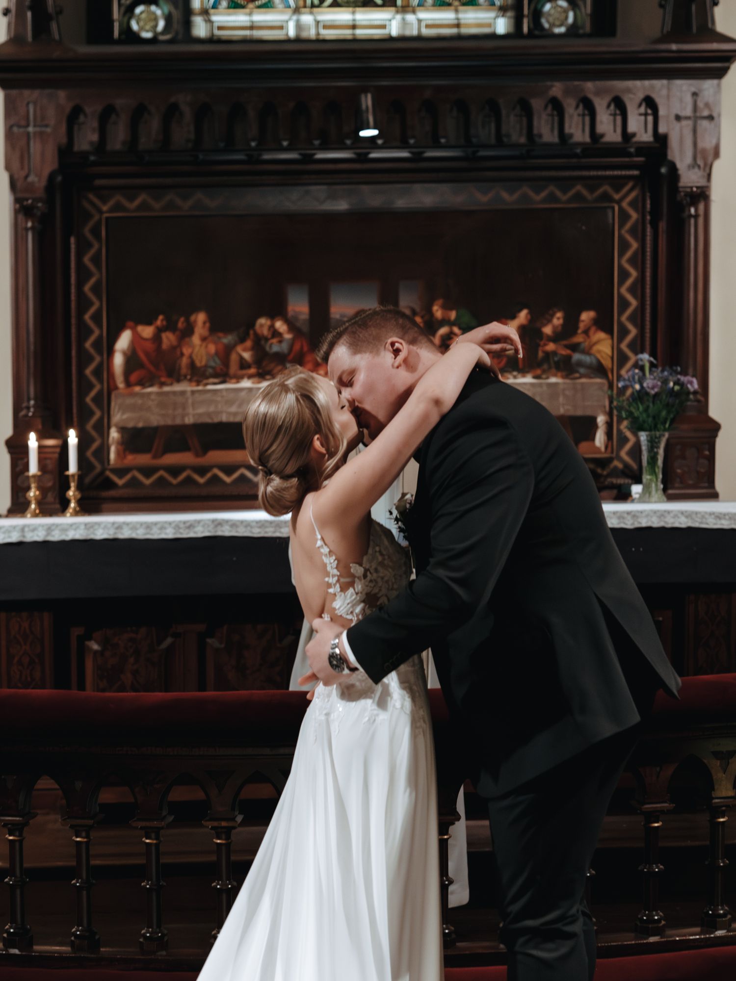 A romantic wedding kiss at the altar with an ornate Last Supper artwork displayed in the background of a church.