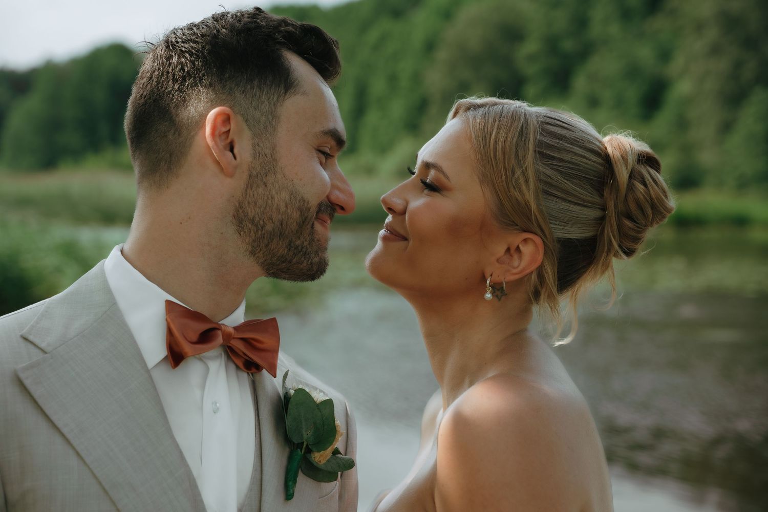 A romantic wedding couple share an intimate moment by a lake, with the groom wearing a gray suit and coral bow tie.