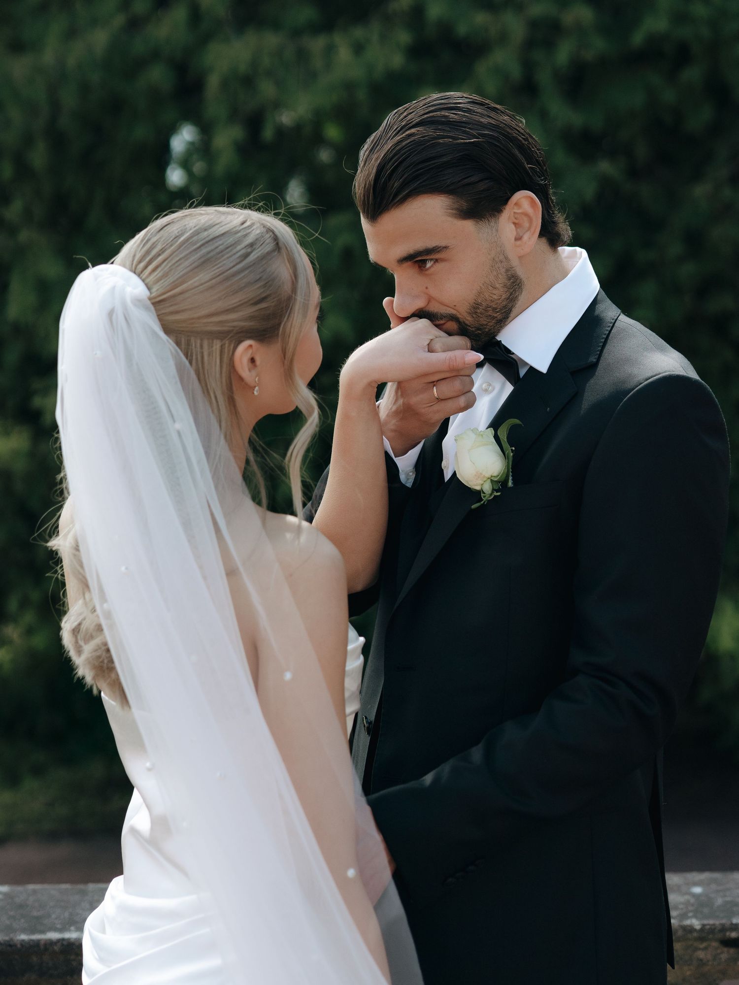 Groom in black tuxedo kissing bride's hand, bride wearing white veil, romantic wedding portrait outdoors.