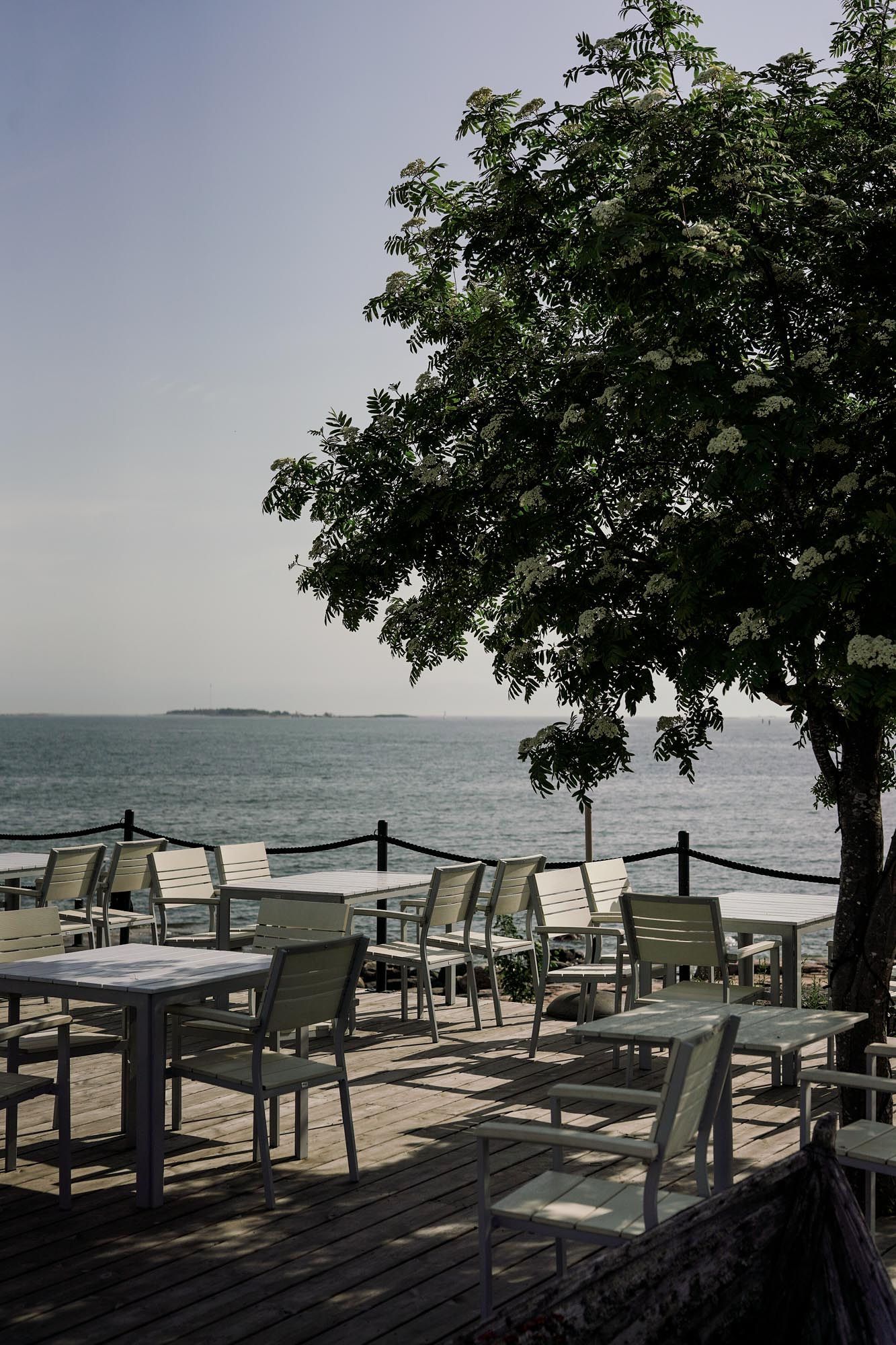 Outdoor waterfront restaurant patio with white chairs and tables on wooden deck beside a large tree overlooking calm sea.