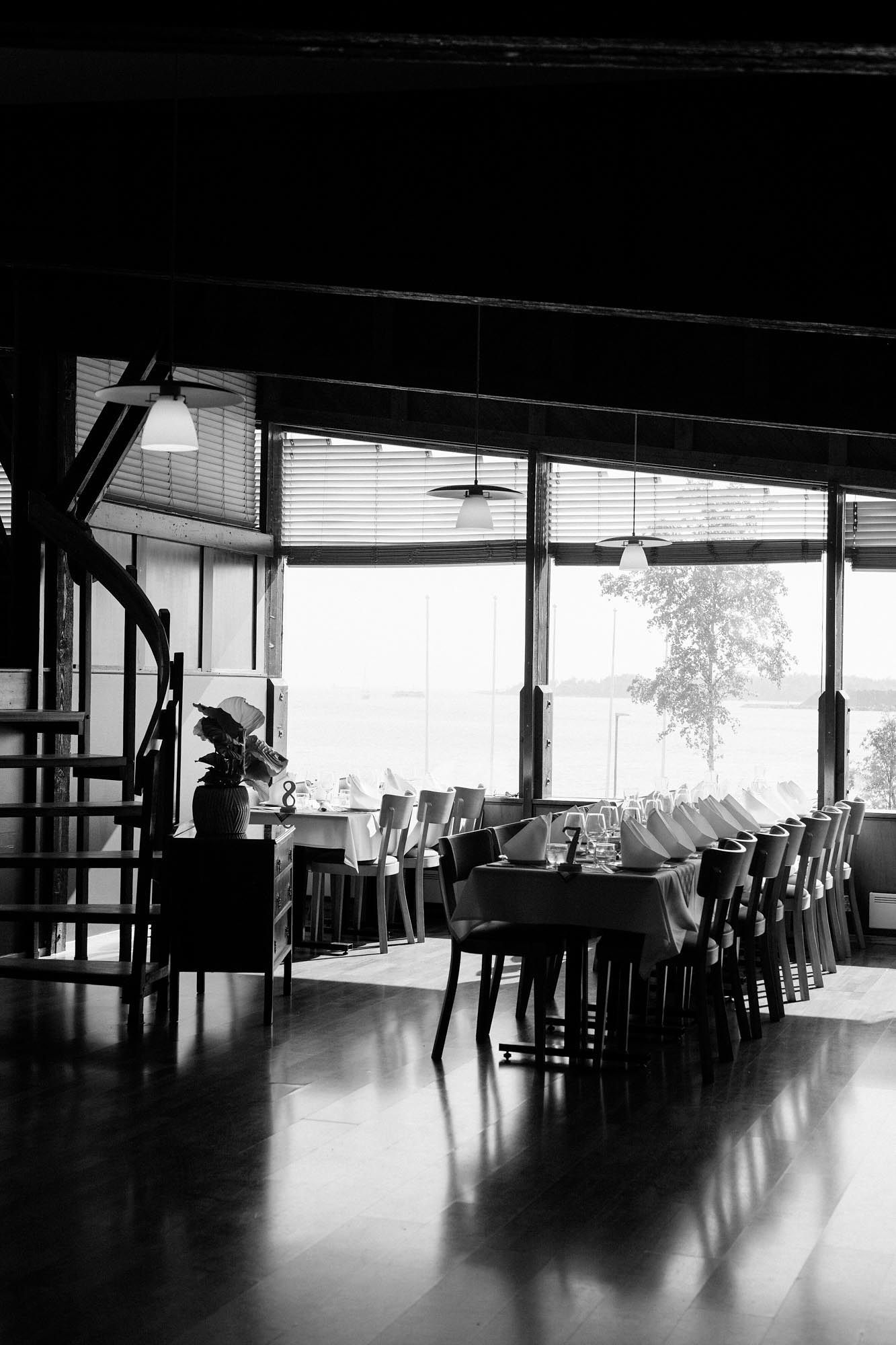 Black and white photo of an elegant restaurant interior with long dining table, chairs, and large windows overlooking nature.