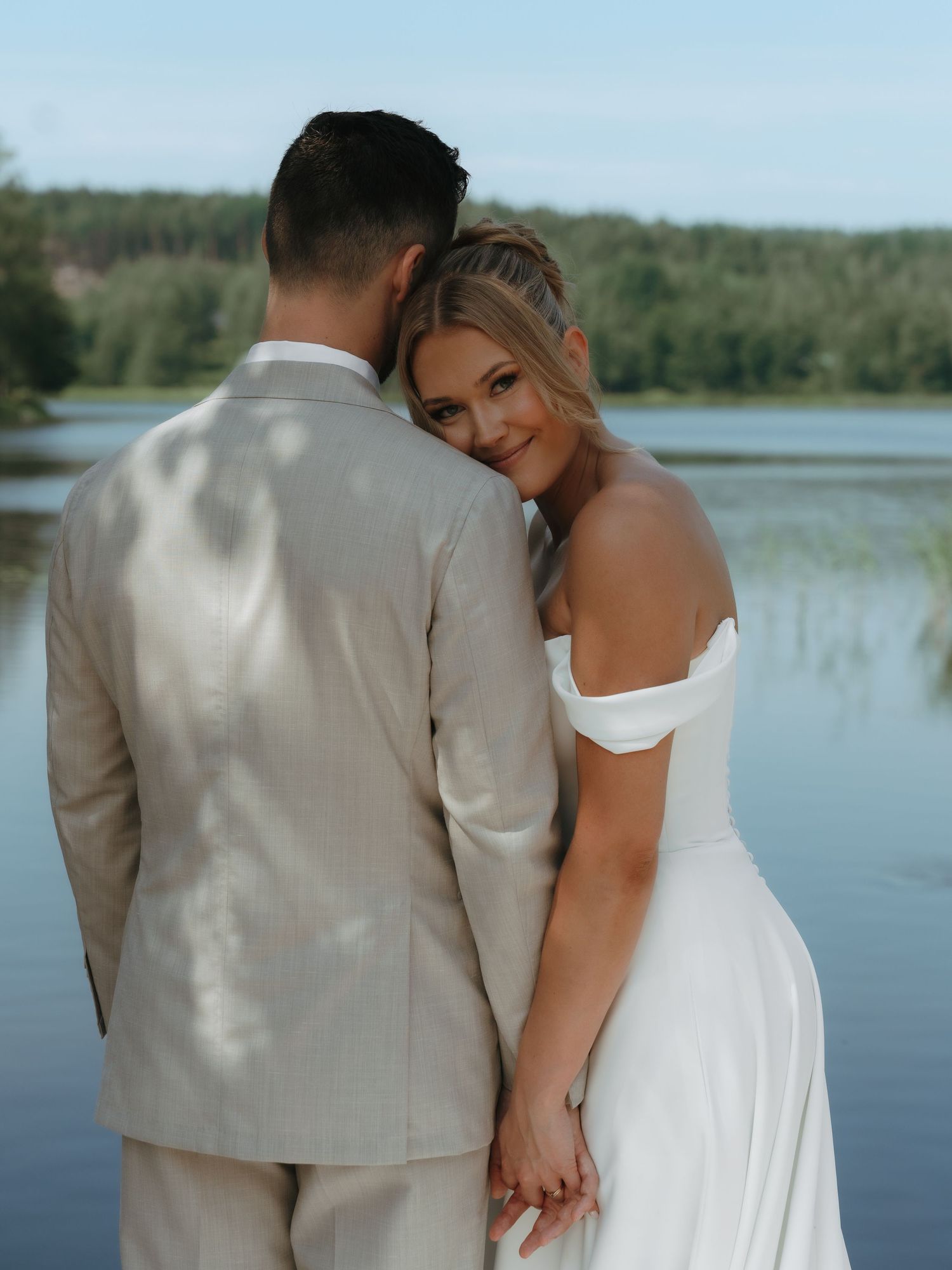 Newlywed couple shares intimate moment in white wedding attire against scenic lakeside backdrop.