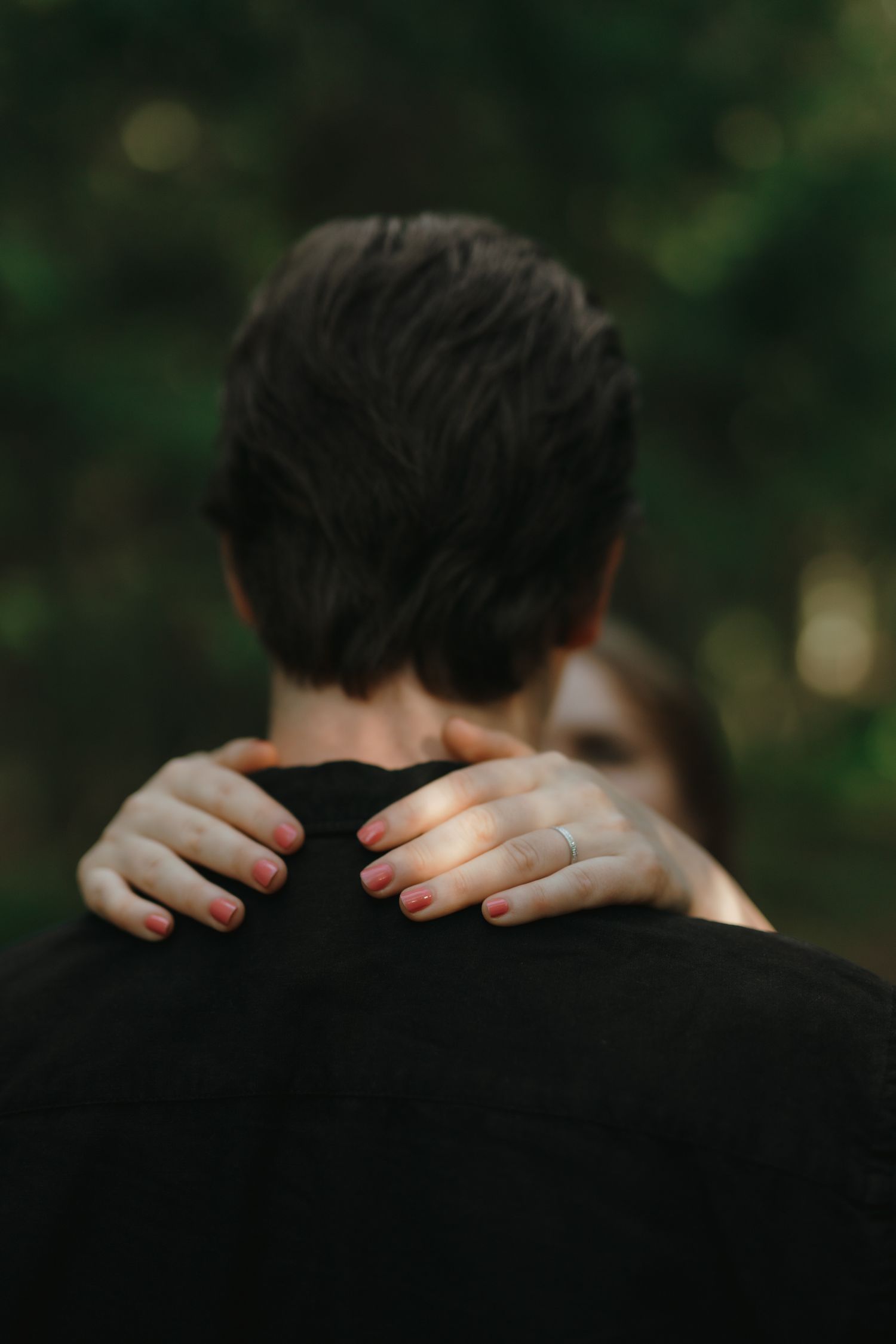Close-up photo of hands with red nail polish embracing from behind against a dark blurred background.