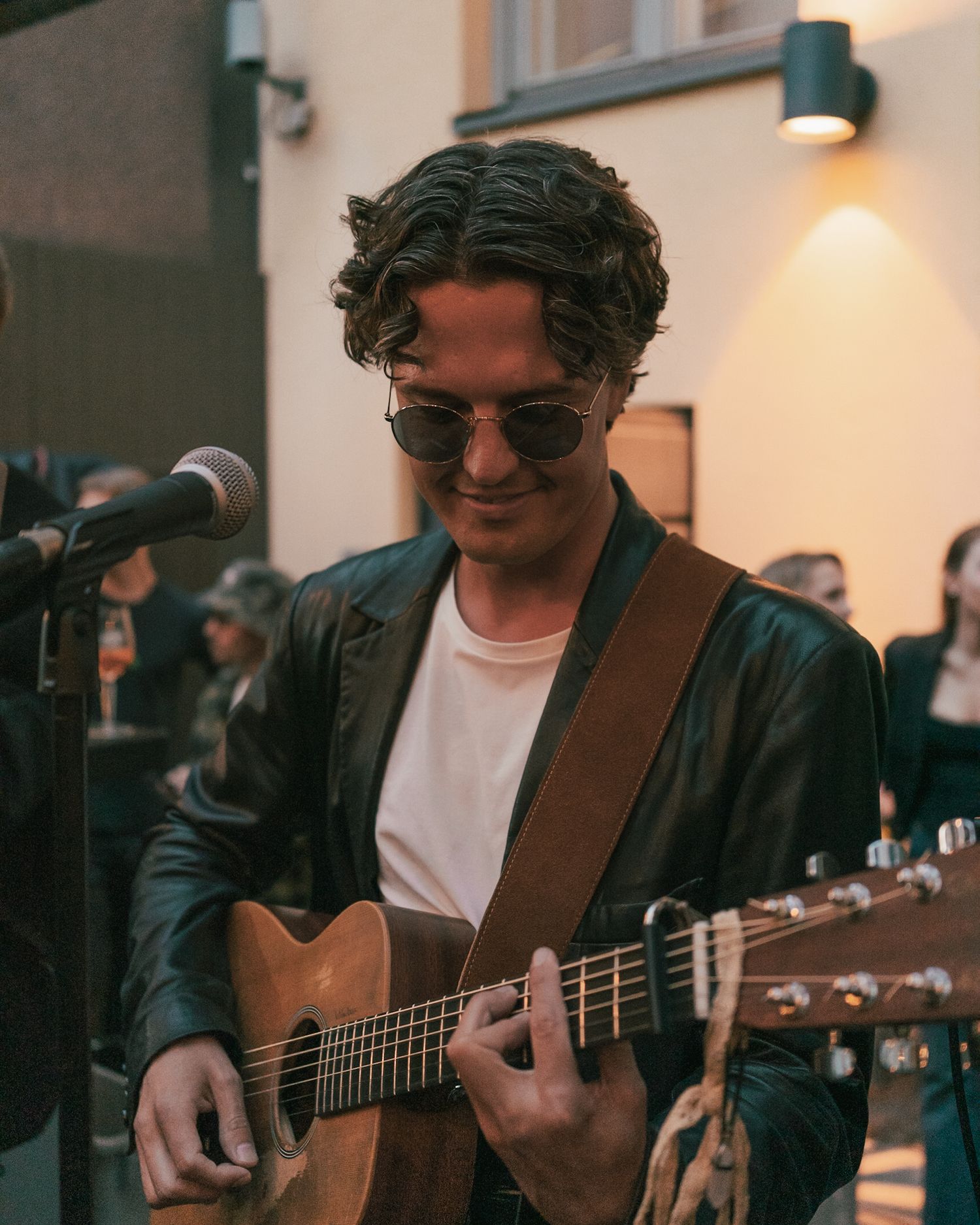 A musician in sunglasses and leather jacket plays acoustic guitar in front of a microphone at an outdoor performance.