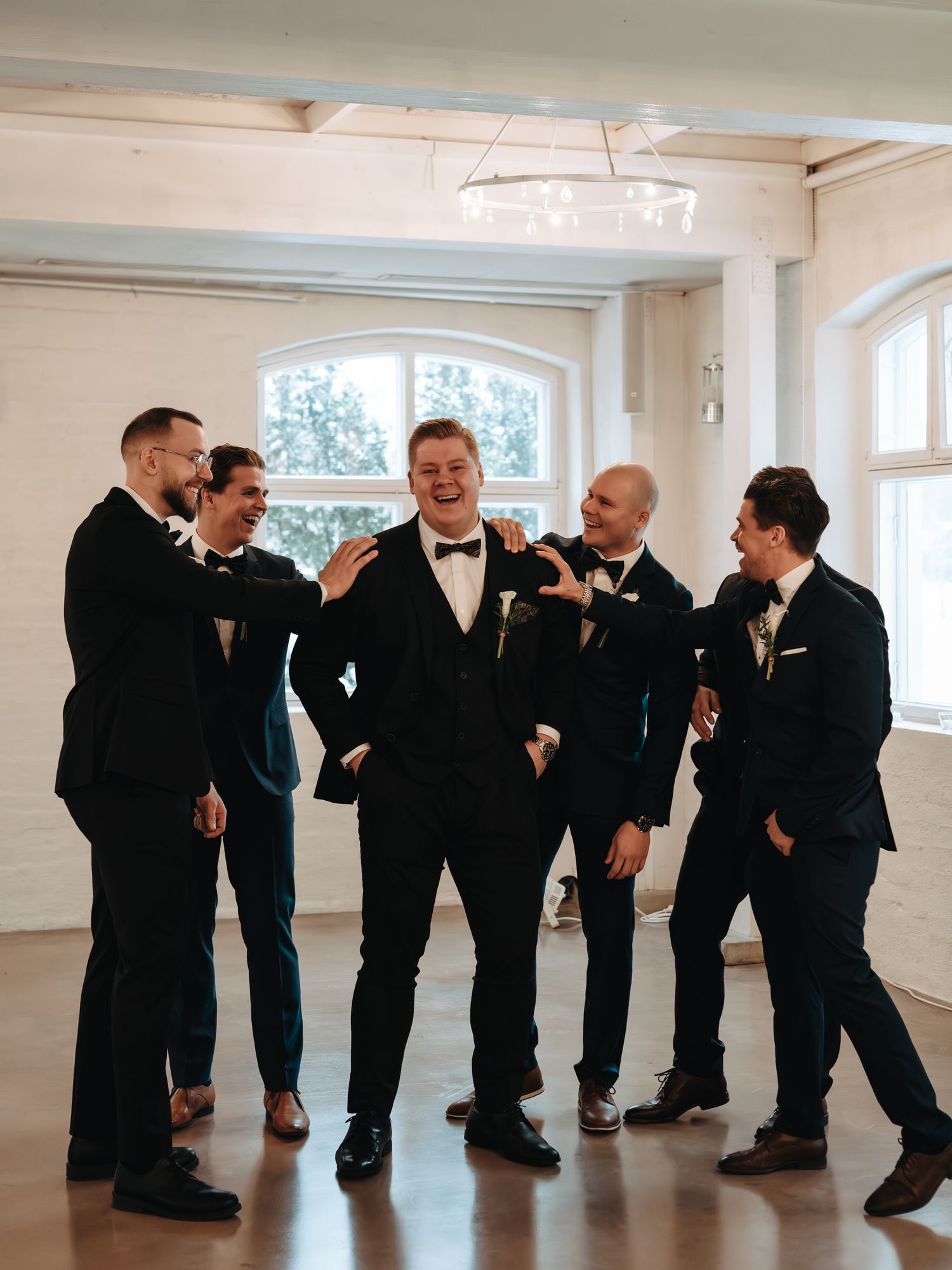 A group of groomsmen in black tuxedos share a candid moment together in a bright indoor space with arched windows.