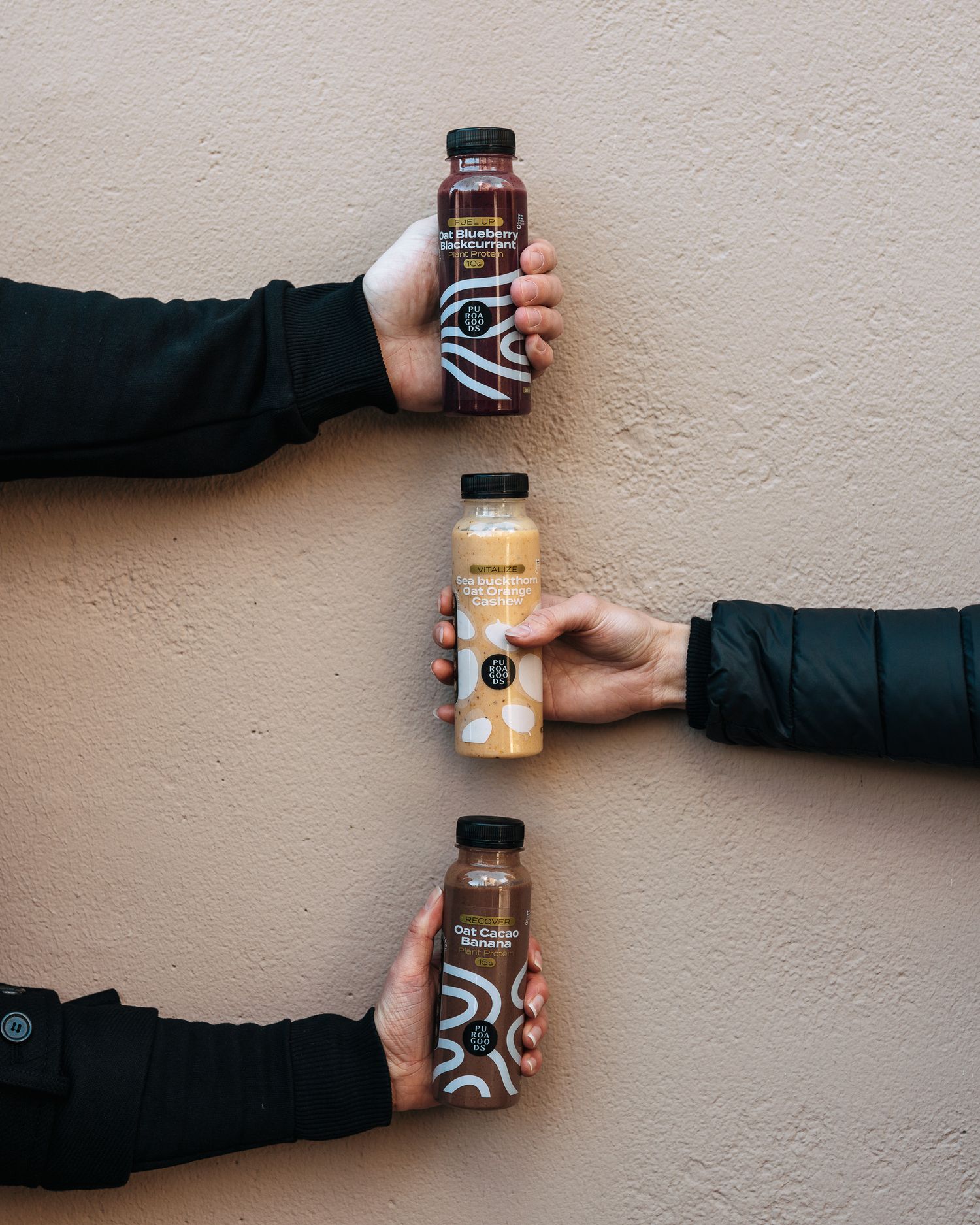 Three hands holding glass bottled beverages with black and white patterned labels against a beige wall.
