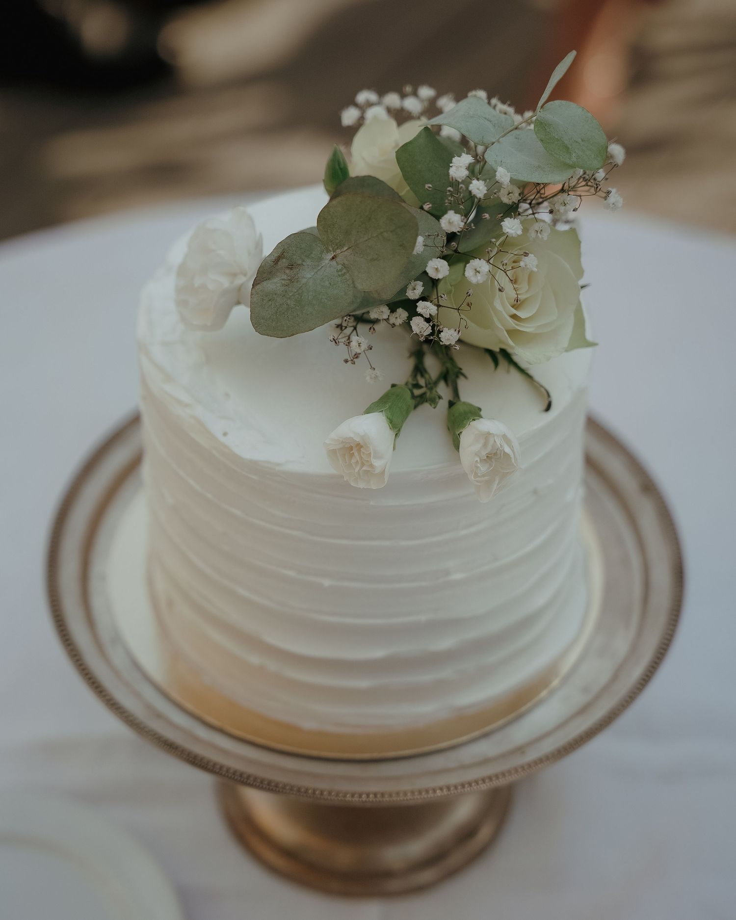 A small white wedding cake with textured frosting decorated with eucalyptus leaves, white roses and baby's breath on a gold stand.