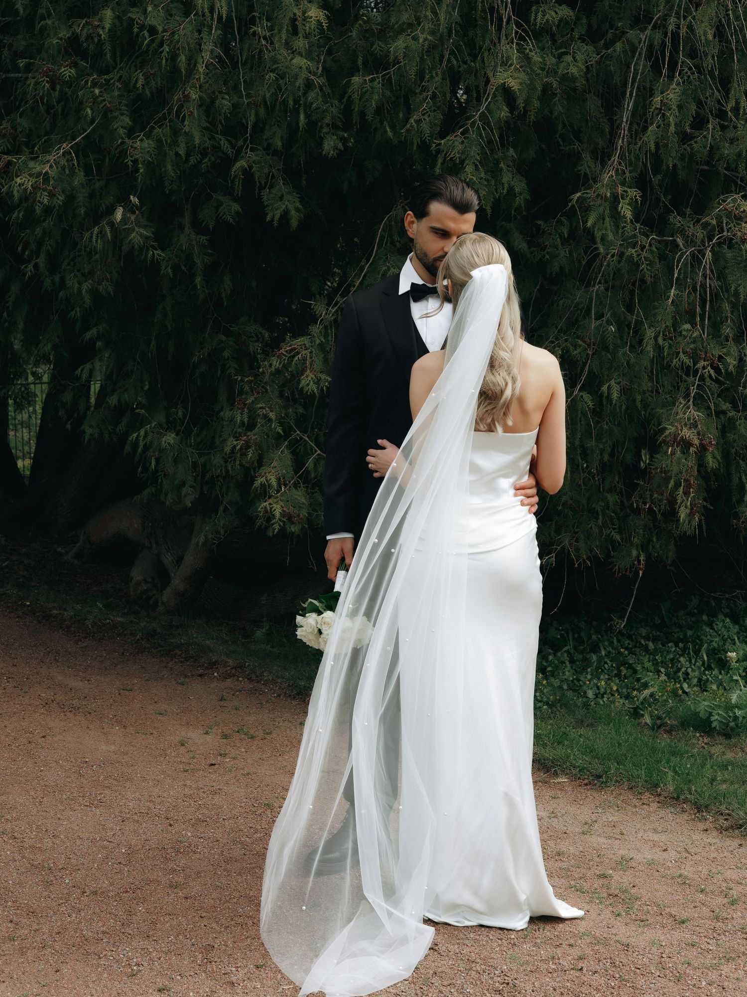 A bride in a white gown and veil holds flowers as a groom in a black tuxedo kisses her near evergreen trees.