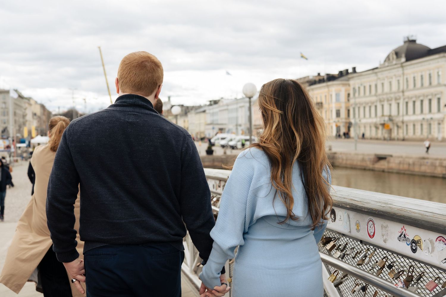 Couple holding hands walking on a bridge by a canal in Helsinki, viewed from behind.