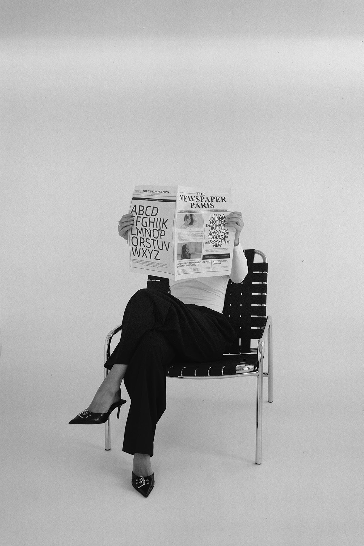 A person in black attire sits on a modern chair reading a newspaper in a minimalist black and white setting.