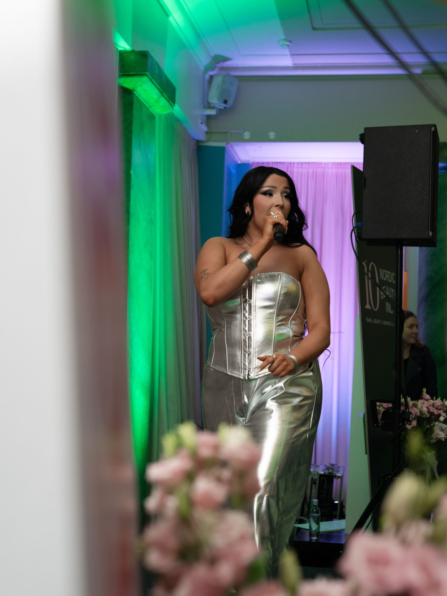 A performer in a metallic silver dress singing on stage with green and purple mood lighting in the background.