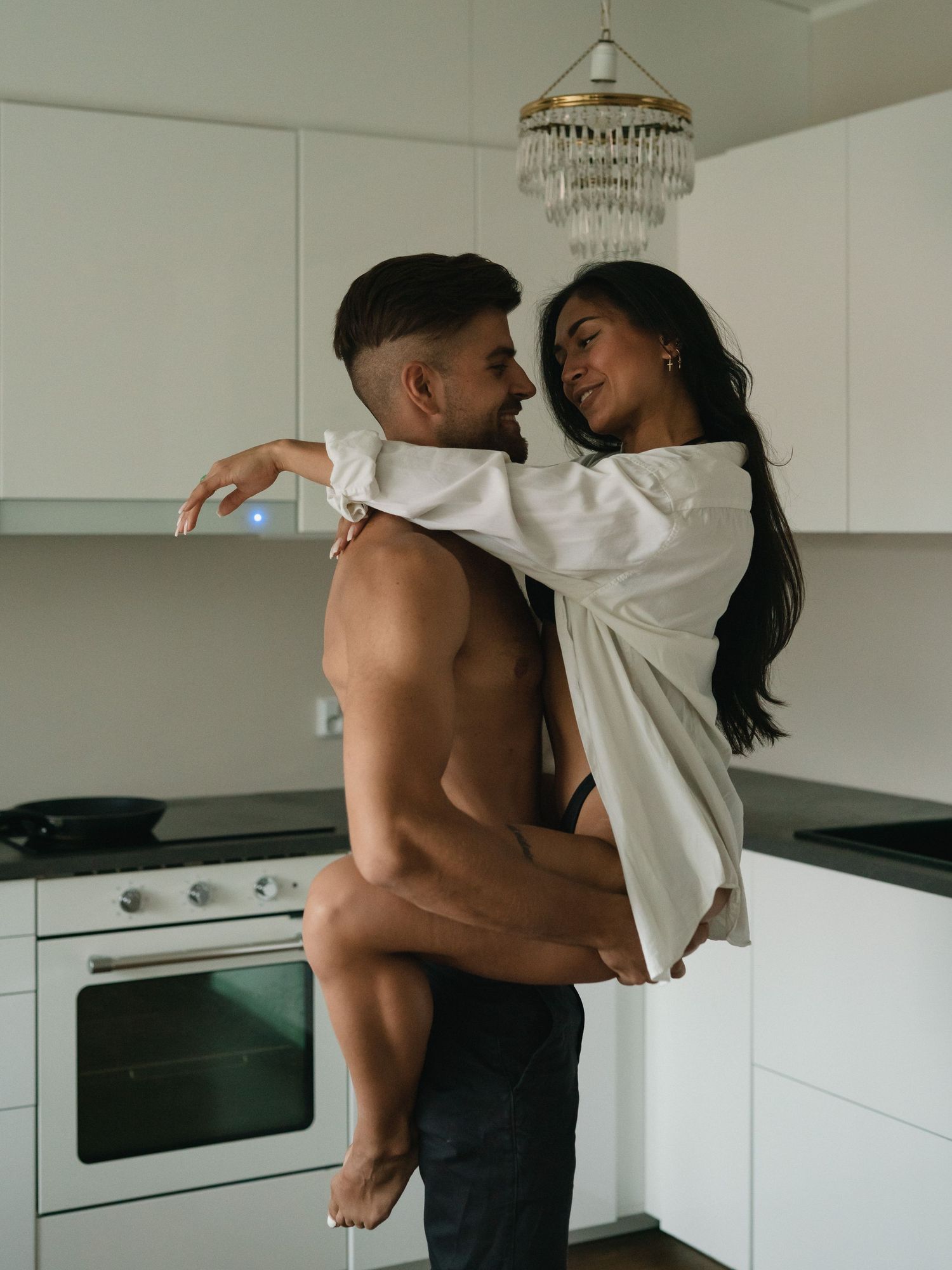 A romantic couple embraces intimately in a modern white kitchen with a crystal chandelier overhead.