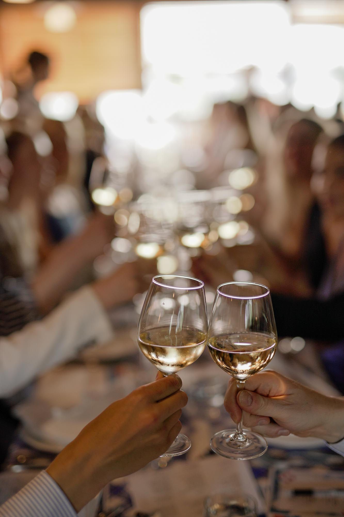 Two hands clinking glasses of white wine at a celebratory dinner party with guests in the background.