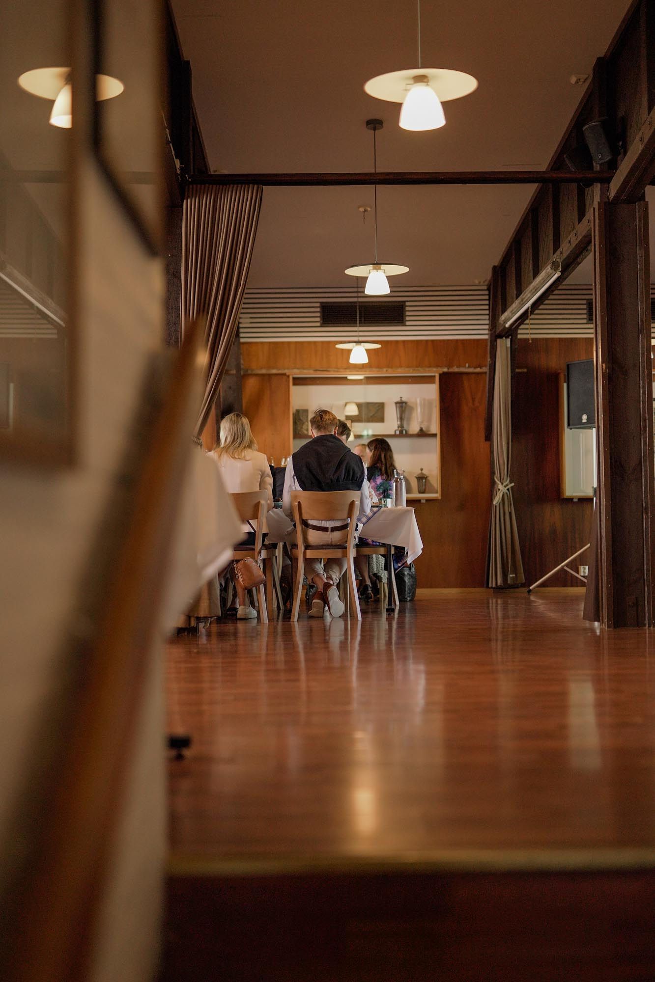 People dining in a warmly lit restaurant with wooden walls, pendant lights, and a long hallway leading to seated guests.