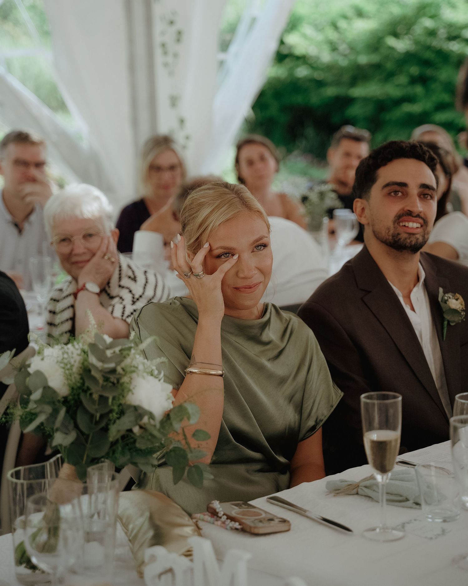 Wedding guests seated at a reception table under a white tent with floral decorations and champagne glasses.