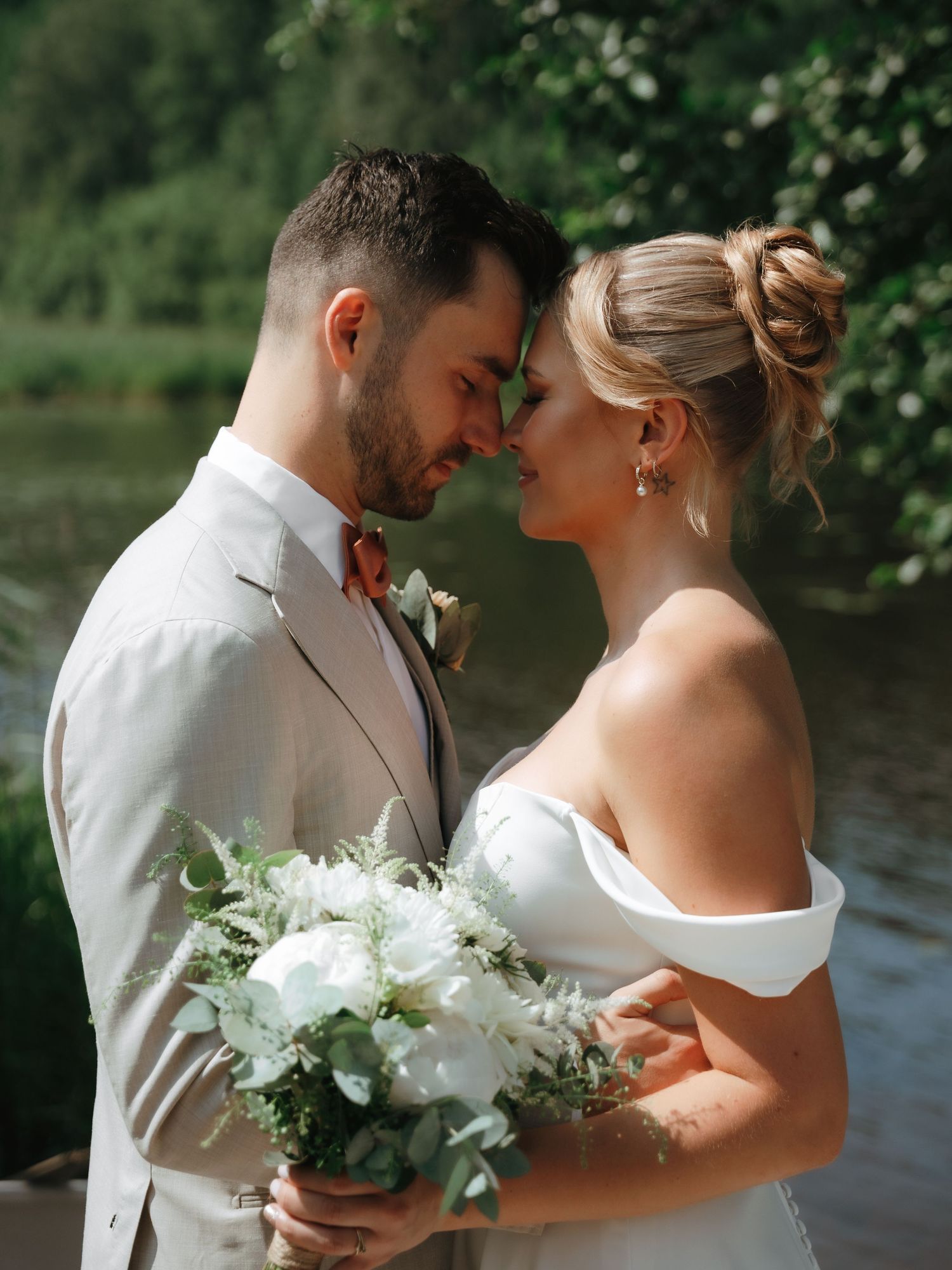 A romantic couple embraces near a lake with white floral bouquet on their wedding day.