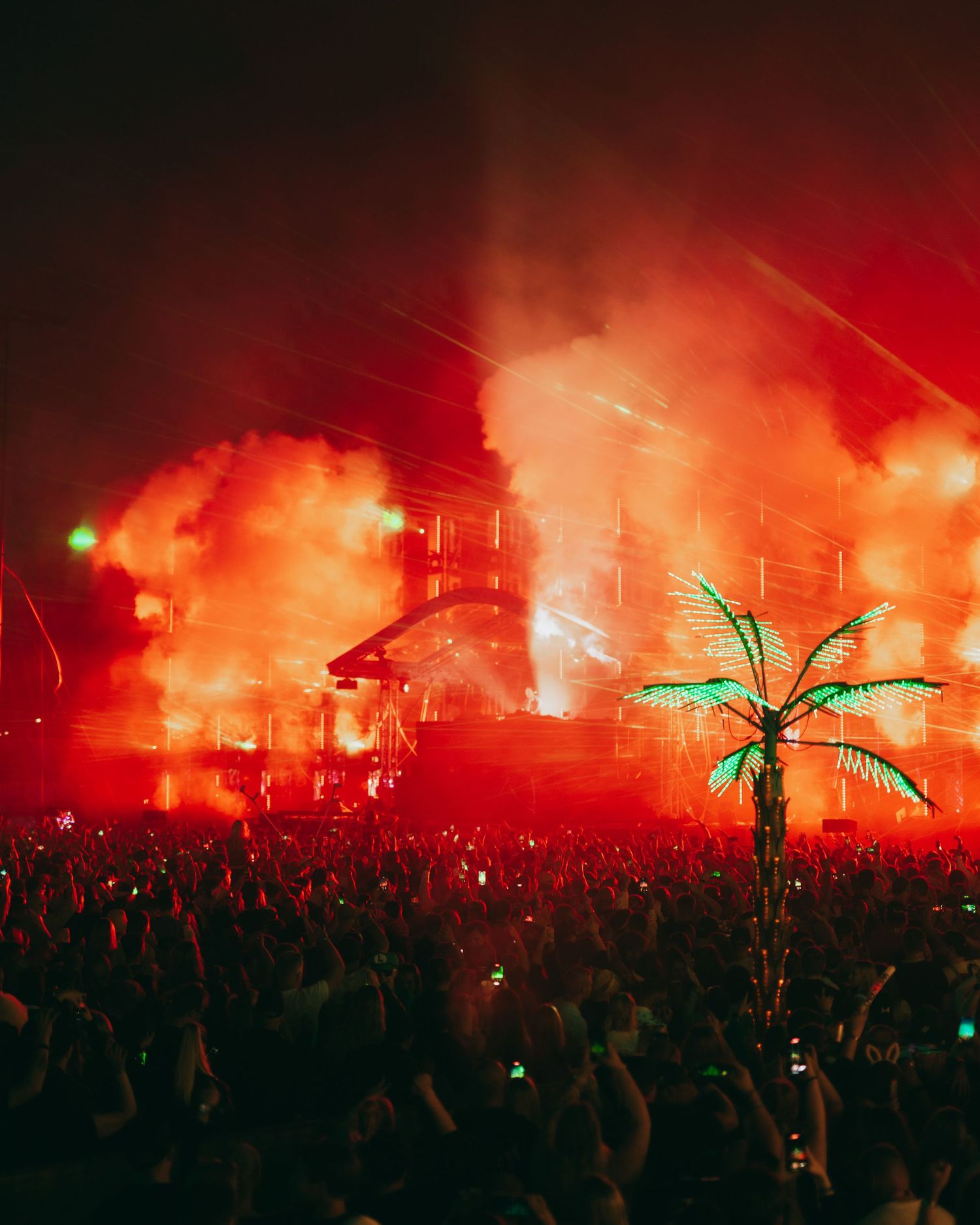 Illuminated concert stage with red smoke effects and neon palm tree decoration amid a large crowd at night.