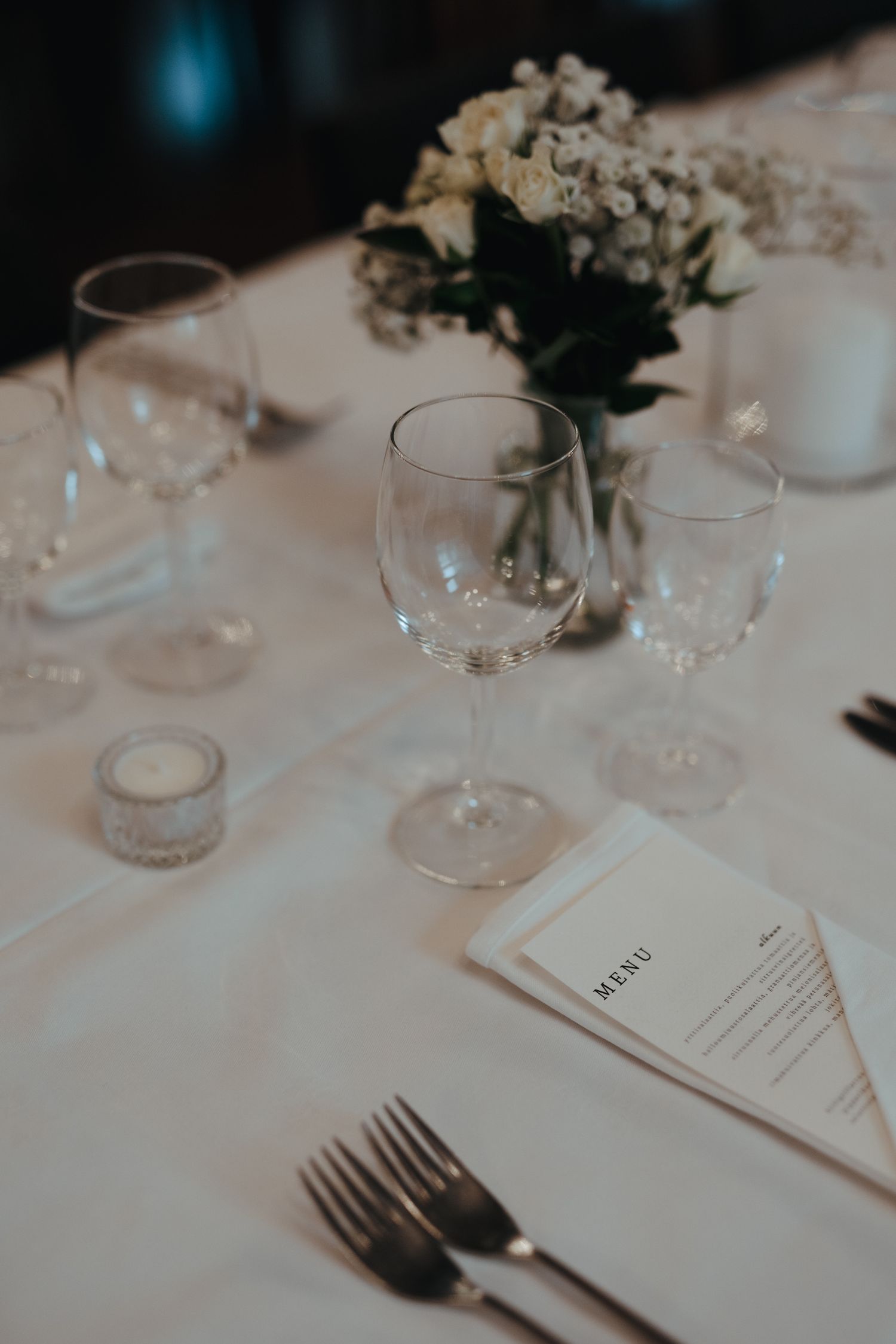 Elegant table setting with wine glasses, white flowers, and menu card on crisp white tablecloth for formal dining.
