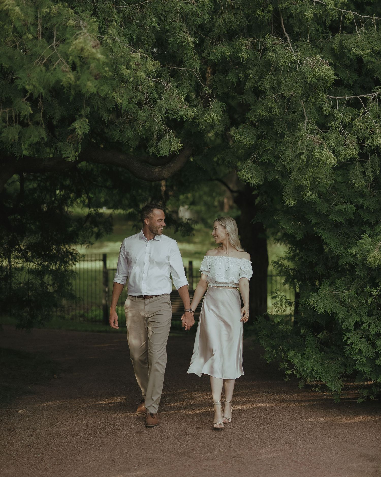 A couple takes a romantic stroll along a tree-lined path in a park at dusk.